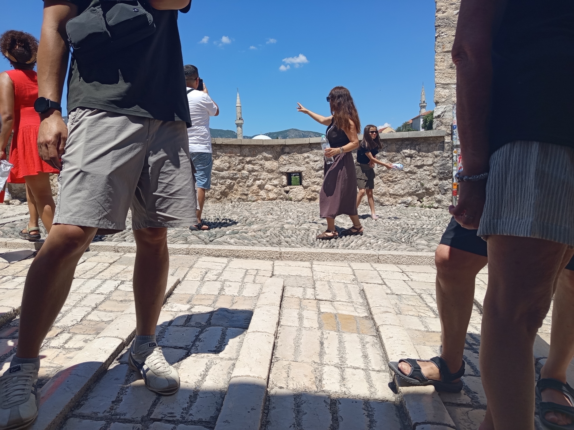 The image shows a sunny day scene with several people near a stone wall in Mostar, Bosnia and Herzegovina. In the foreground, a person wearing grey shorts and white sneakers is partially visible from the waist down. To the left, a woman in a red dress walks away from the camera, and another person in a white t-shirt and blue shorts is visible in the background. In the middle ground, a woman in a dark dress points towards a minaret in the distance, holding a bottle of water. Another woman in a patterned skirt walks behind her. The background features a stone wall, cobblestone ground, a minaret, and a dome, characteristic of the Old Town of Mostar.