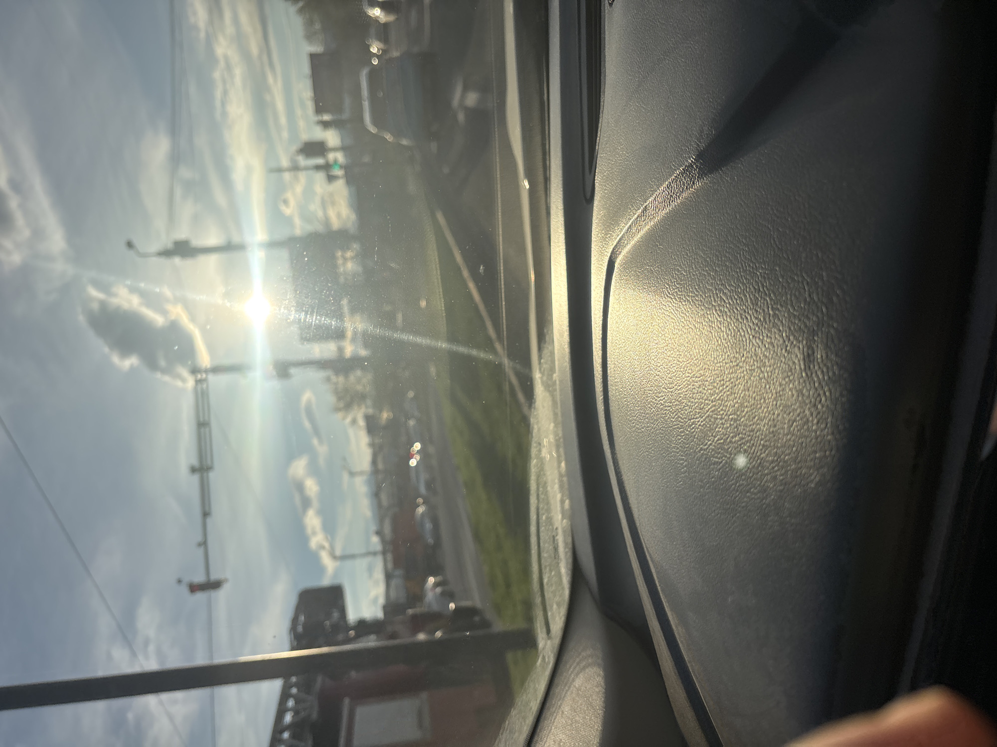 The image shows a view from inside a car, looking out the front windshield. The sun is shining brightly, causing glare on the glass. Outside the car, there's a street scene with traffic lights and buildings. There appears to be a grassy area along the side of the road. Based on the architectural style and the presence of Georgian license plates visible, the location is likely Tbilisi, Georgia. The interior of the car features a textured dashboard, and the focus is split between the dashboard and the outside view. A finger is also visible on the lower right corner of the frame.