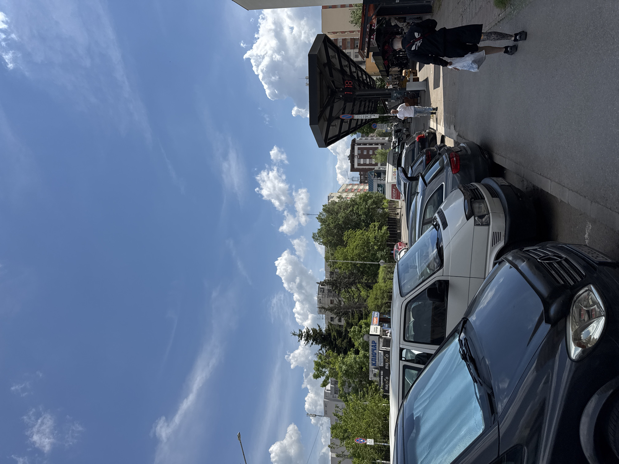 The image captures a street scene in Sofia, Bulgaria. Several parked cars line the right side of the frame, including a dark-colored Mercedes-Benz in the foreground and a white van behind it. In the background, trees and apartment buildings are visible, typical of urban areas in Sofia. Pedestrians are walking on the sidewalk. The sky above is blue with fluffy white clouds. A bus stop is seen in the background.