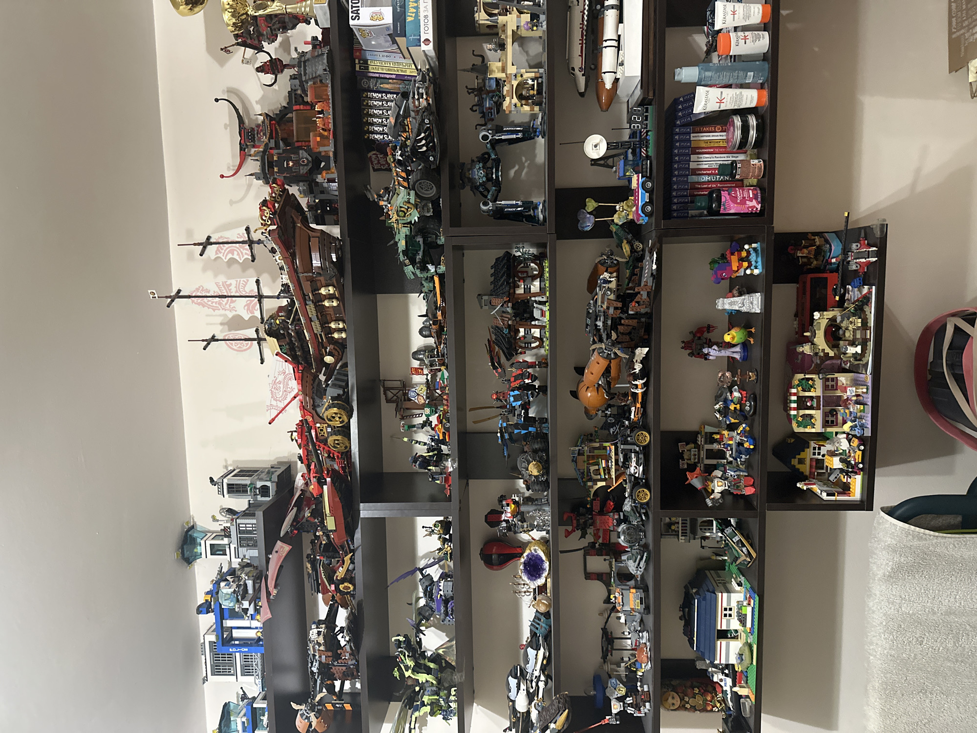 This image captures a multi-tiered shelving unit against a light-colored wall in a room located in the city of Unknown, Unknown. The dark-colored shelving unit contains six horizontal shelves filled predominantly with various LEGO sets and other collectibles.
On the top shelf, from left to right, are multiple white and blue LEGO City Police sets, including a multi-story police station, a helicopter, and a small control tower. To their right, a large brown LEGO ship with three masts and red sails is displayed. Adjacent to the ship is a dark grey LEGO fortress-like structure featuring red horns. Further right, a dark grey and black LEGO mechanical figure stands next to a golden trophy cup.
The second shelf displays a large green and black LEGO dragon figure on the left. To its right, a purple LEGO flying vehicle is positioned. In the center, a large green and black LEGO monster truck-like vehicle is visible. Further right, a stack of books, including several volumes of "Demon Slayer" manga, are arranged vertically.
The third shelf features several white, grey, and blue LEGO vehicles, some resembling aircraft. A purple geode is centrally placed. To the right, a large black and blue LEGO bipedal mech figure is displayed next to a light-colored LEGO castle-like structure. A white and orange LEGO space shuttle set is partially visible on this shelf.
On the fourth shelf, a small wooden Russian nesting doll is on the far left. A white and blue LEGO house with a dark blue roof is situated in the center-left. Multiple small LEGO vehicles, including a police car, are placed around it. To the right, a yellow-roofed LEGO building with green awnings and various LEGO minifigures is displayed.
The fifth shelf contains a tan and brown LEGO structure resembling a desert building with minifigures and a vehicle. A red LEGO double-decker bus model is also present. To the right, a collection of personal care products, including multiple Kérastase branded bottles (white, blue, orange, green), a dark pink bottle, and a small brown bottle, are arranged. Behind these products, a stack of PlayStation 4 game cases is visible. A digital clock displays "3:21."
The bottom shelf features several smaller LEGO vehicles and architectural models. In the bottom left foreground, a white textured towel or blanket is draped over an unseen object. A portion of a red and blue chair is visible in the bottom right foreground.