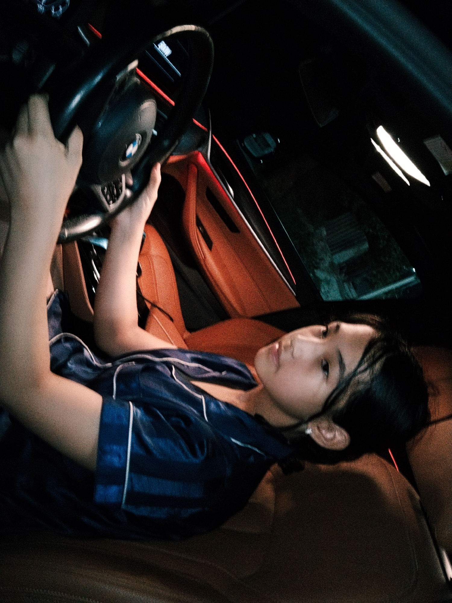 A young girl is seated in the driver's seat of a car, holding the steering wheel. She is wearing navy blue silk pajamas with white trim. The car's interior features tan leather seats and ambient red lighting along the dashboard and door panels. The girl is looking to her right, with her head tilted back slightly. The scene is dark, suggesting it is nighttime, and the car appears to be stationary. There is no indication of the location being Koçkor-Ata, Kyrgyzstan.