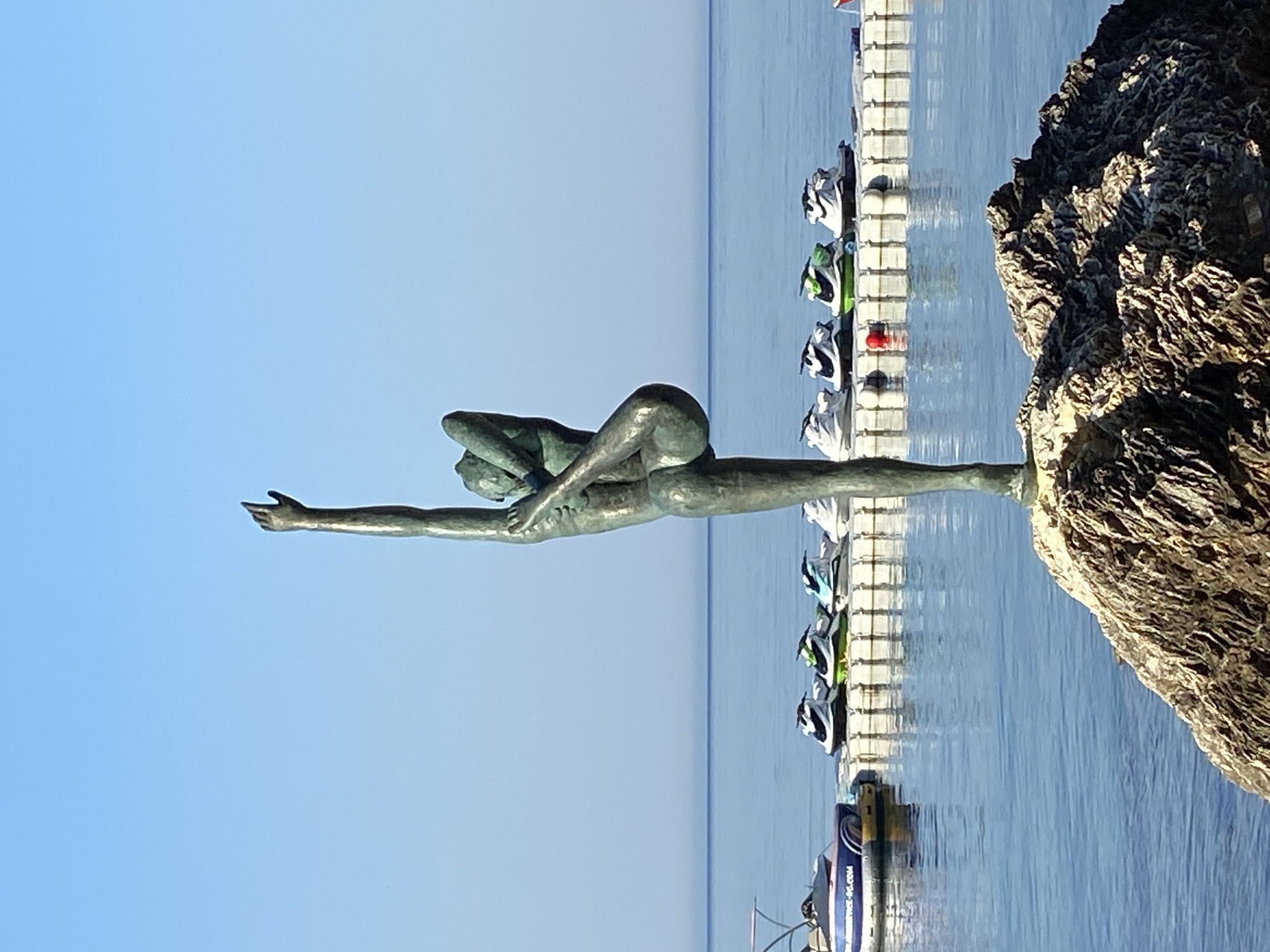 The image captures a bronze sculpture of a person in a graceful, yoga-like pose, standing on a rocky outcrop. The sculpture is positioned in front of a calm blue sea, with a clear, bright sky overhead. In the background, a white floating dock is visible, lined with several jet skis. To the left of the dock, a smaller boat with a canopy is partially submerged in the water. The overall scene suggests a scenic coastal location. This sculpture is located on the promenade of Budva, Montenegro, a popular tourist destination known for its beautiful beaches and vibrant atmosphere.