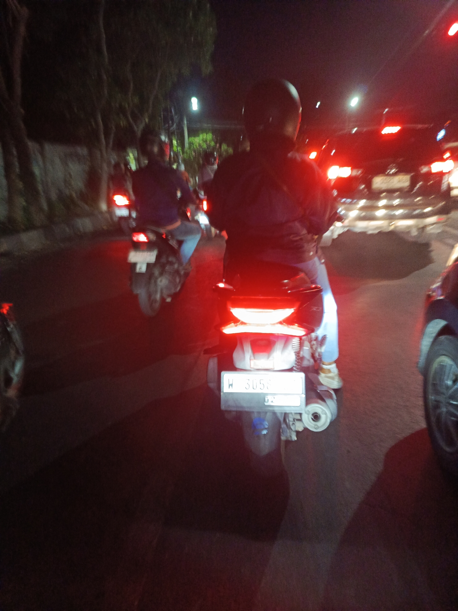 The image depicts a nighttime scene of traffic on a street. In the foreground, a scooter is stopped, with its brake lights illuminated. The scooter has a license plate that reads "W 3058". A person wearing a dark jacket and jeans is seated on the scooter, facing away from the camera. They are wearing a helmet. Behind the scooter, other vehicles are visible, also with their lights on, indicating a traffic jam. Trees and streetlights line the side of the road. The overall impression is one of a typical urban traffic situation at night.