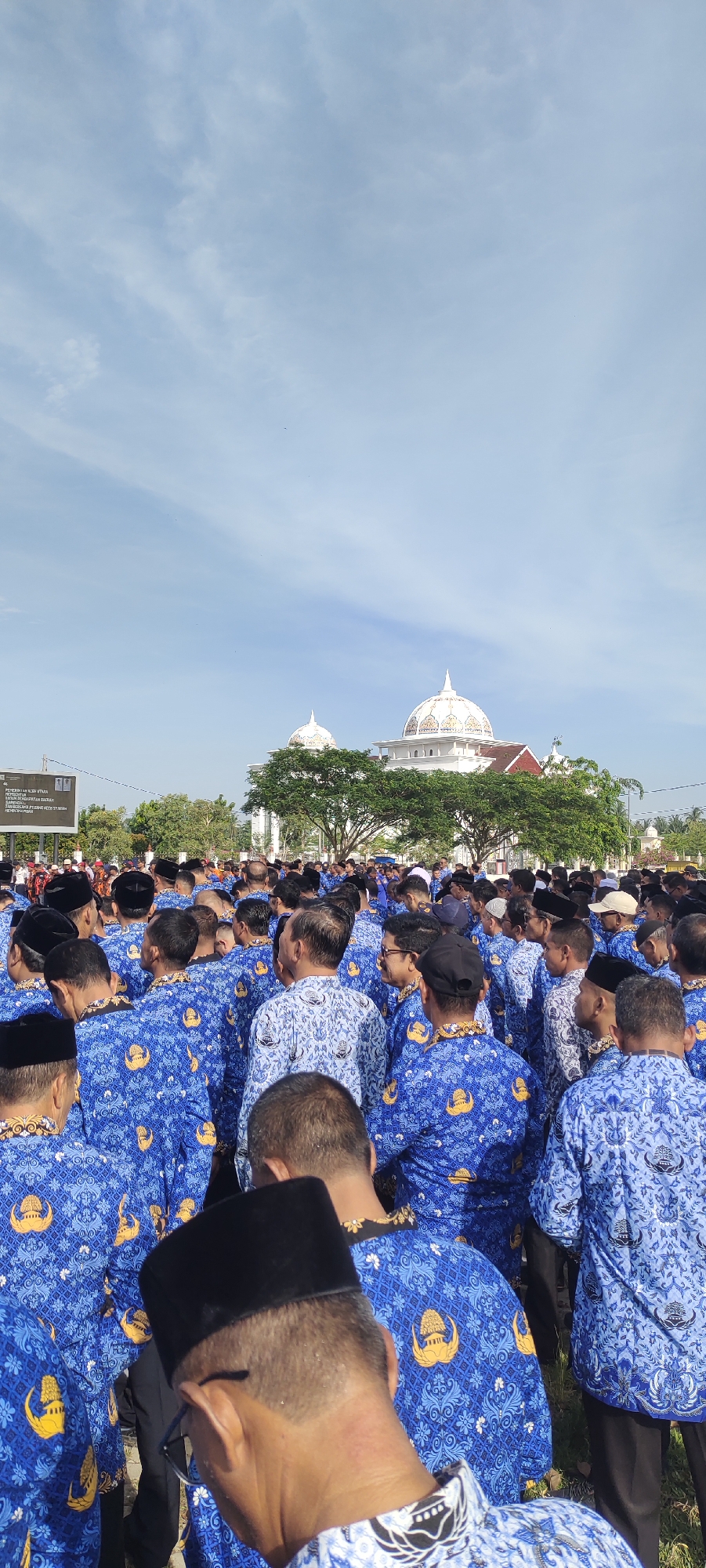 A ground-level, eye-level, full outdoor shot shows a large group of men in blue patterned shirts with black caps, standing closely together in front of a large white building with a dome, under a light blue sky.
The men are predominantly seen from the back, with their heads and shoulders visible. They are wearing blue shirts with a repeating pattern and gold-colored crests, and many are wearing traditional black caps.
In the background, a large white building with a prominent dome is visible. It is framed by green trees.
The sky is light blue with some wispy clouds. In the mid-ground, a dark sign board is visible with Indonesian text, as well as some other bystanders. The location is outdoors in Meunasahaluedrienlhoksukon Barat, Indonesia.