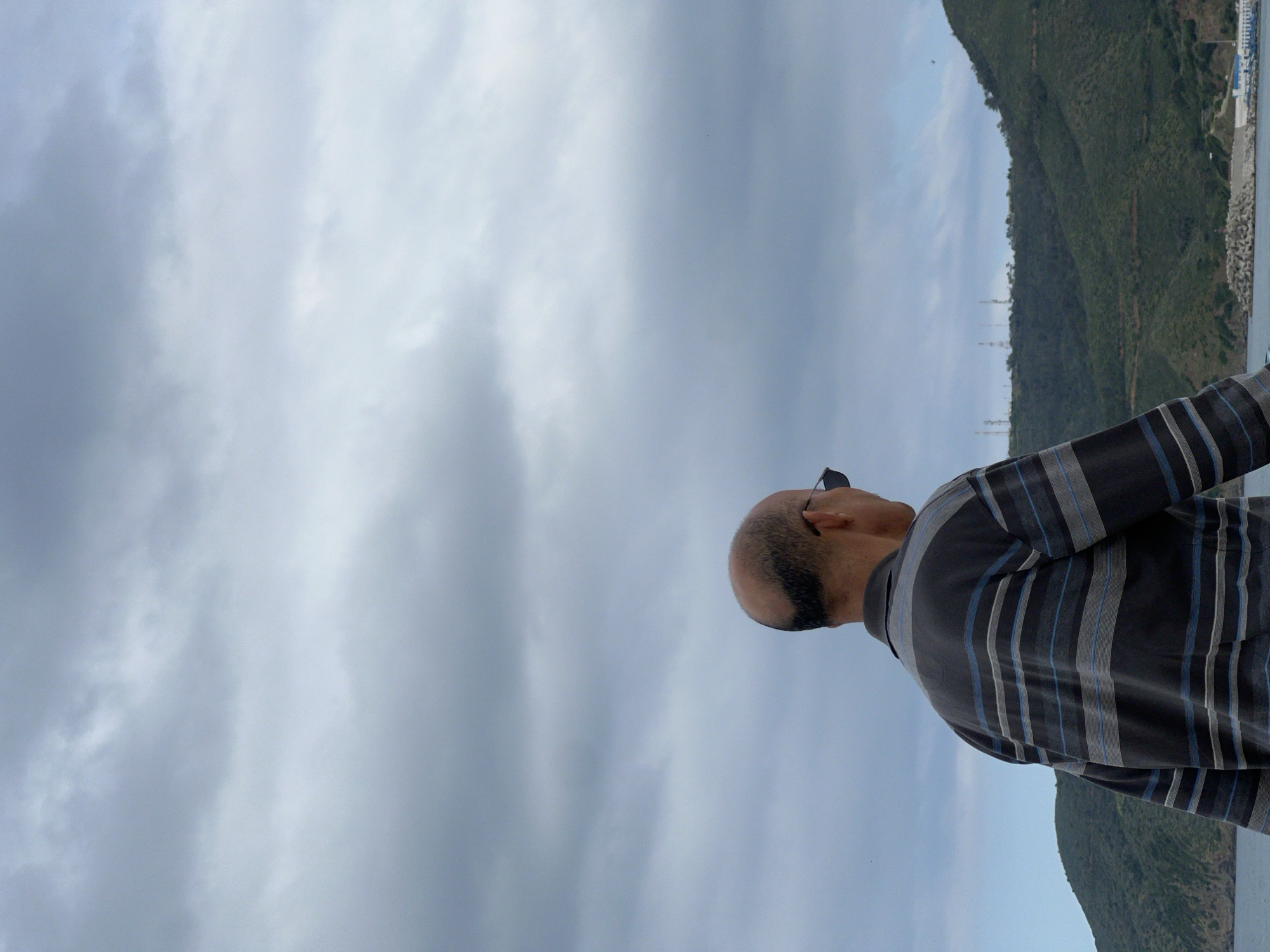 A man stands outdoors, facing a landscape of hills and water, in M'Diq, Morocco. He is bald with dark hair on the sides and back of his head, wearing sunglasses and a striped shirt. The sky is overcast with cloudy formations. In the background, a green, mountainous terrain slopes down toward the water, alongside a constructed wall with white blocks and structures.