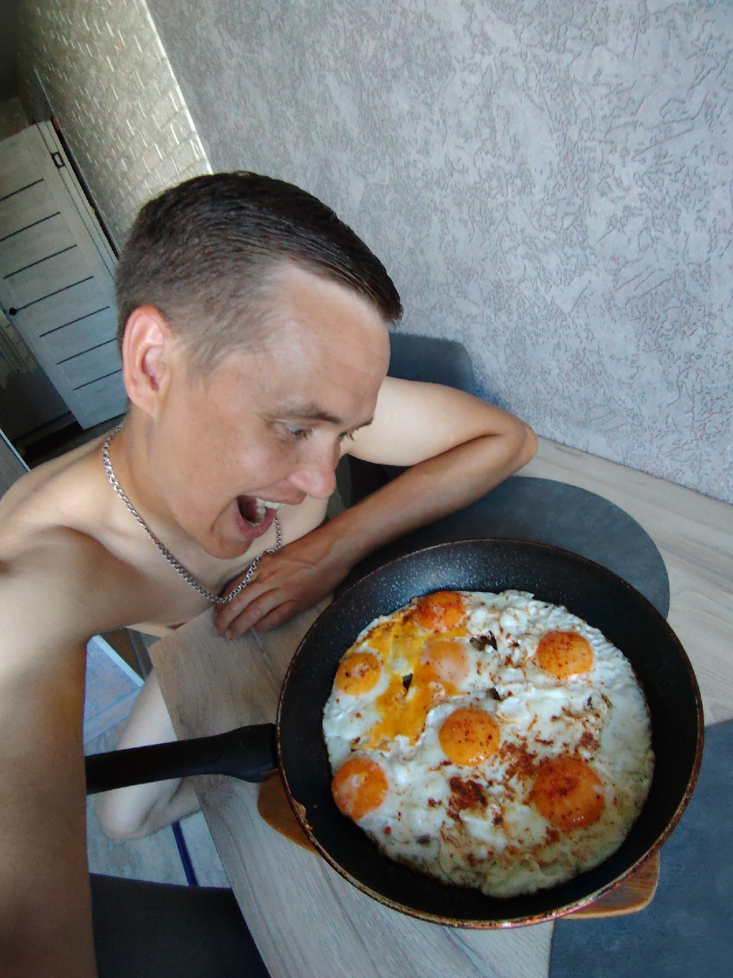 A man with short, dark hair is shown from the chest up, leaning towards a black frying pan filled with fried eggs. The man is wearing a silver chain necklace. His mouth is open in an exaggerated expression. The frying pan is on a wooden surface, likely a table. The background features a textured wall and a partially visible closet or door. The setting appears to be indoors, likely a residential kitchen or dining area. The image is taken from a slightly elevated perspective, looking down at the man and the frying pan.