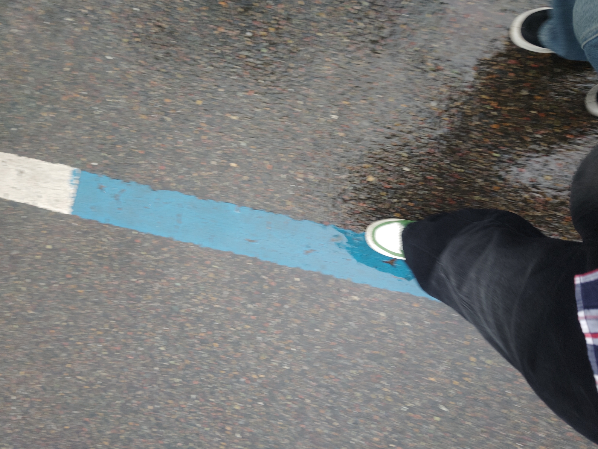 The image shows a person standing on wet asphalt in Chongqing High-tech Zone, China. The ground is marked with a blue and white stripe. The person is wearing dark pants and a shoe that partially covers the blue stripe. Another person wearing jeans and sneakers is visible in the upper right corner of the image. The ground appears wet, possibly after rain.