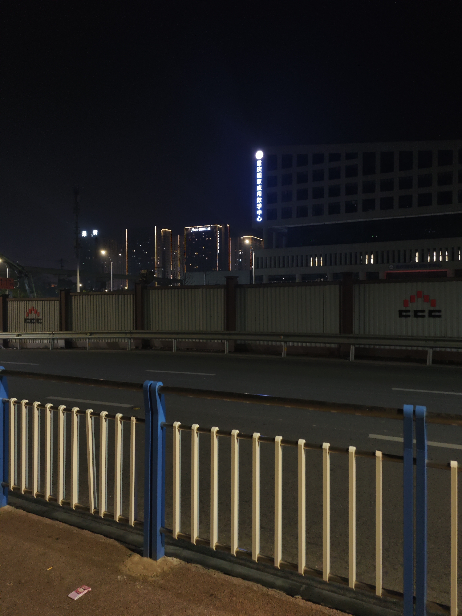 The photograph depicts a nighttime street scene in Chongqing High-tech Zone, China. In the foreground is a blue and white metal railing. Beyond the railing is a two-lane road. A barrier lines the opposite side of the road, with the CCC logo visible on it. Behind the barrier are several illuminated buildings, including a large, box-shaped building with lit Chinese characters vertically aligned on its side. Additional buildings with vertical lines of light are also present in the background. The sky is dark.