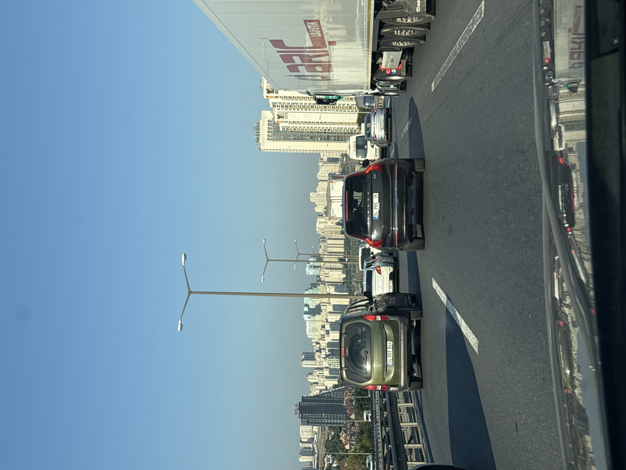 A daytime, eye-level, vertical shot captures a traffic jam on a highway with the cityscape of Esenyurt, Turkey, in the background. The sky is clear and blue. Several lanes of traffic are visible, with multiple cars and a large truck in motion. The foreground shows the reflection of the cityscape and traffic lights on the hood of a vehicle. In the middle ground, a green Peugeot wagon with license plate "34 HDY 944" is in the left lane, and a black Volvo SUV with license plate "34 PHS 35" is in the right lane. A white semi-truck with "BRIC LOGISTIK" written on its side is in the far right lane. The background reveals a dense urban environment with numerous tall buildings. Streetlights line the highway.