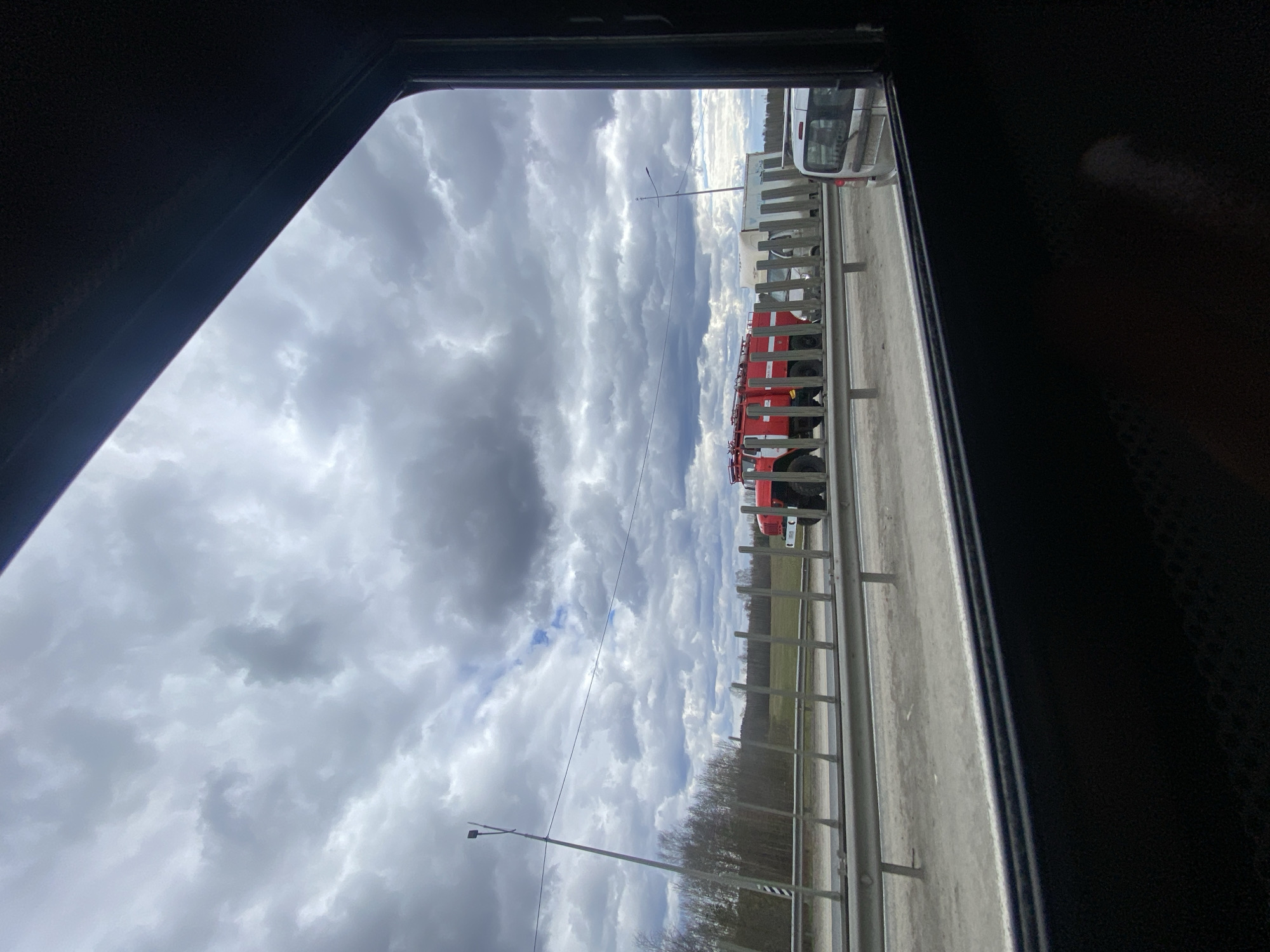 The image shows a view through a window, possibly of a vehicle, looking out onto a cloudy sky. A line of red fire trucks is parked on a road alongside a metal guardrail. Beyond the guardrail, there's a strip of green grass and a line of trees. The location appears to be on a roadside, potentially a highway or a main road in the city of Unknown, Unknown. There are streetlights visible.