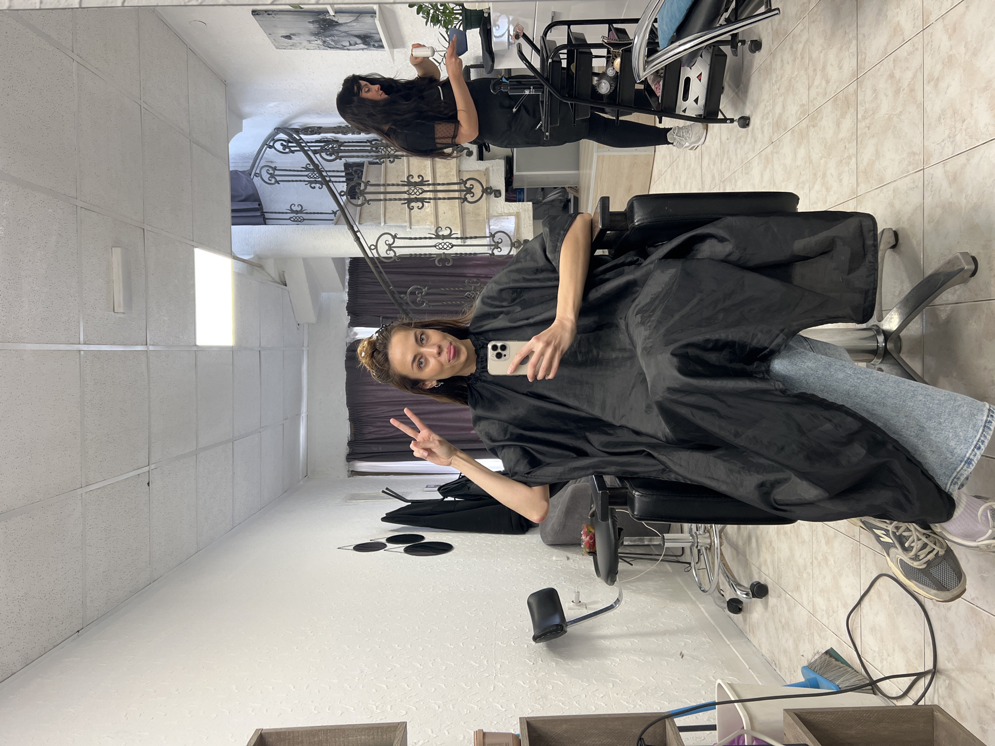 The image shows a woman sitting in a salon chair, covered in a black cape, holding a phone, and making a peace sign. Her jeans and sneakers are visible. A second woman, presumably a hairdresser, is standing in the background near a trolley. The reflection in the mirror shows a staircase with ornate iron railings. The location appears to be a salon, possibly in Bar, Montenegro, judging by the interior design and architectural details visible in the reflection. The floor is tiled. The walls are white. There is a window with drawn blinds behind the woman in the chair. The setting is indoors.