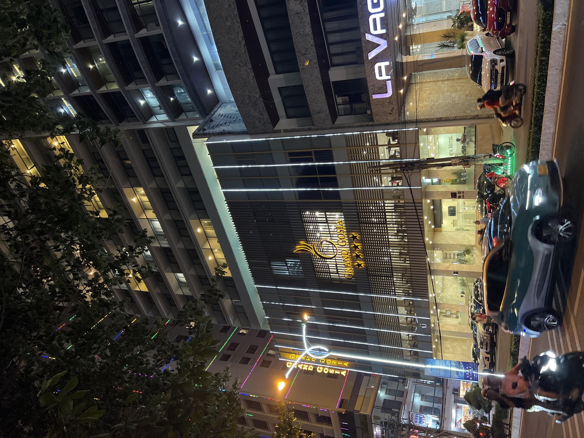 This image captures a nighttime street scene in front of the "GRAND GOSIA" hotel in Nha Trang, Vietnam. The hotel building is brightly illuminated with vertical white lights and features signage with the hotel's name and a four-star rating. Adjacent to it, another building displays a neon sign that reads "LA VAGA." The street is busy with traffic, including a scooter with two passengers in the foreground, a teal car in motion, and several parked cars and a white SUV. Pedestrians are also visible on the sidewalk and in the hotel's lobby, which is transparent and shows tables and chairs. Trees with visible foliage hang over the scene, and colorful string lights are draped among them. The overall atmosphere is lively and urban, typical of a city at night.