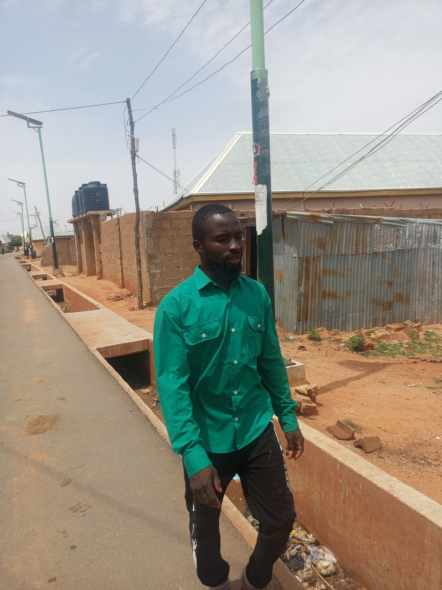 A man stands in the city of Tempure, Nigeria. He wears a green button-down shirt and black pants with a white stripe. He has a dark beard. He is standing in the street next to a concrete drainage ditch filled with trash. Behind him is a building with a corrugated metal wall and a white roof. Power lines and utility poles are visible. Buildings line the street in the background. The sky is light blue.