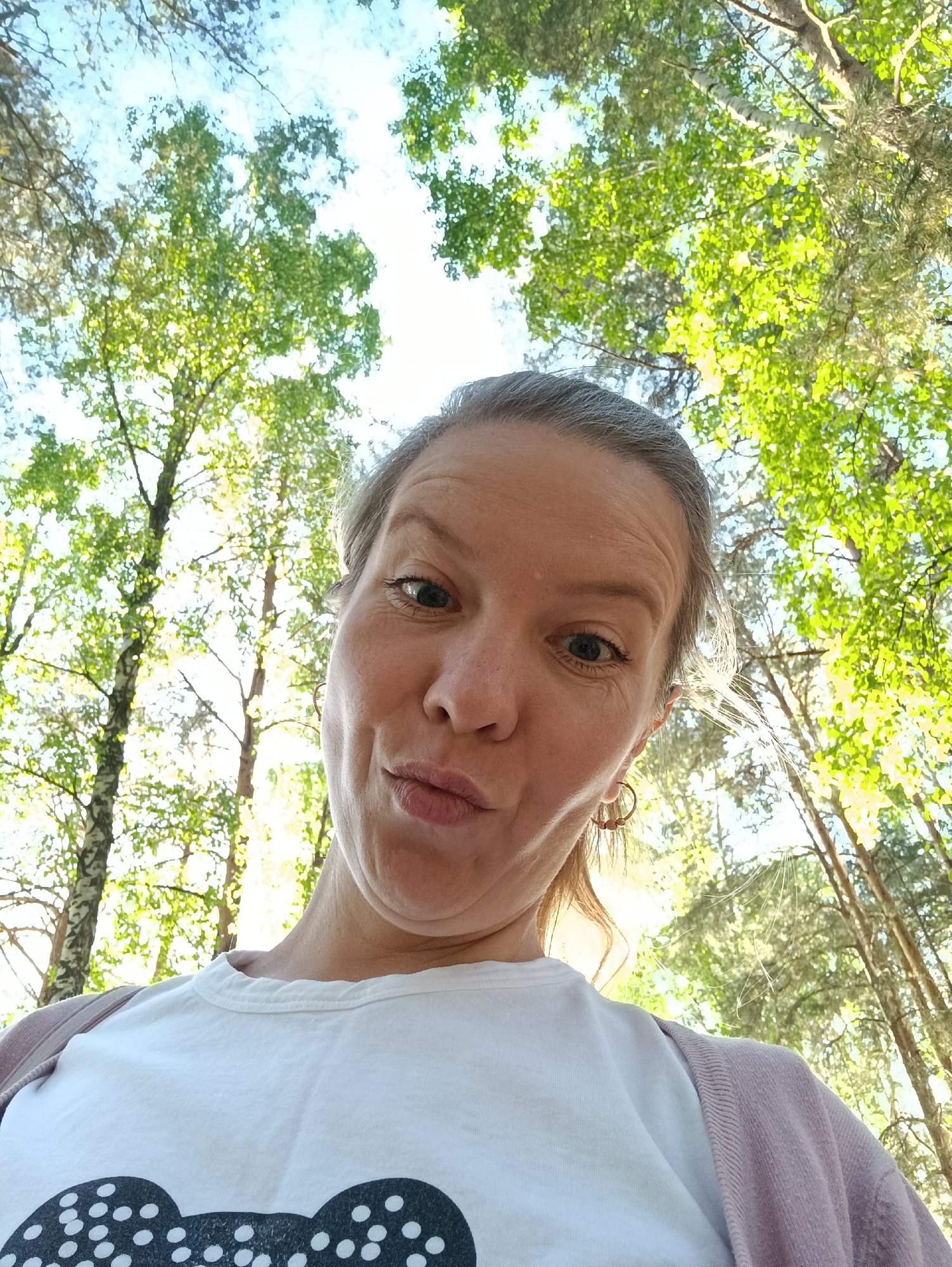 A selfie of a fair-skinned woman with graying hair pulled back into a low bun, taken from a low angle in an outdoor setting, likely in the city of Chaikovsky, Russia. The woman is wearing a white t-shirt with a graphic print on the front and a light pink cardigan. She has earrings and is making a pouting face. The background consists of trees with green foliage and a partly cloudy sky. The trees appear to be birch trees based on their bark.