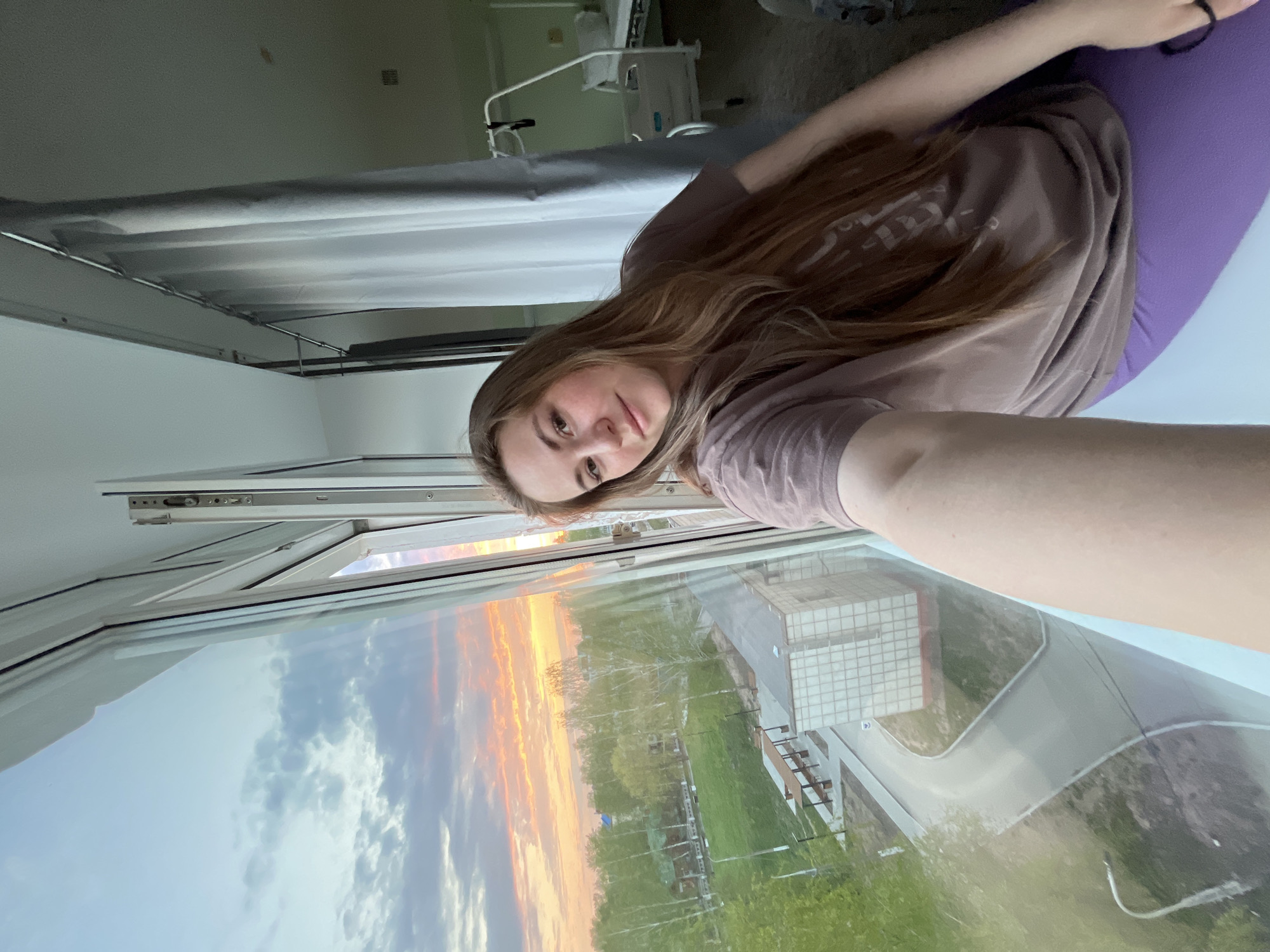 A young woman with long brown hair is positioned in front of a large window displaying a sunset over Chaikovsky, Russia. She is wearing a brown t-shirt and sitting on a purple object. The window provides a view of a building with a grid-like facade, green spaces, a road, and trees outside. Inside, the setting includes a bed, possibly in a hospital or care facility. The photograph appears to be taken with a selfie perspective, with the woman's arm extending towards the camera.