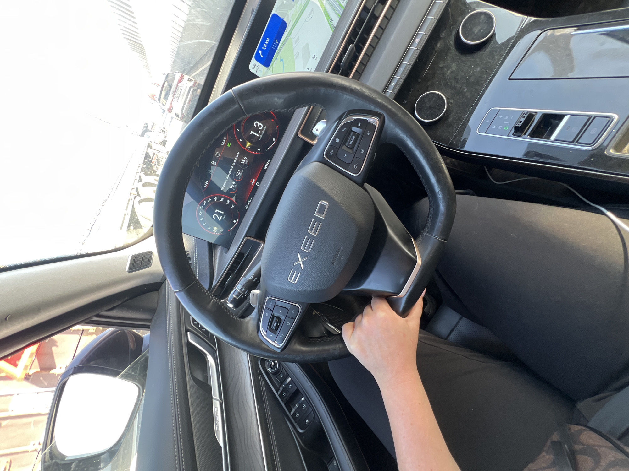 Here is a description of the image:
The image shows the interior of a car, with the focus on the steering wheel and dashboard. The steering wheel is black and has the brand "EXEED" prominently displayed in the center. A hand is visible holding the steering wheel. The dashboard features a digital display showing various information such as speed, fuel consumption, and time. A navigation map is visible on the dashboard's screen. The car's gear shift is in the drive position. The environment suggests the vehicle is in motion. Based on the navigation screen showing the "КАНТЕМИРОВ" (Kantemirov) street, the car is located in Moscow, Russia. The person is wearing black clothing.