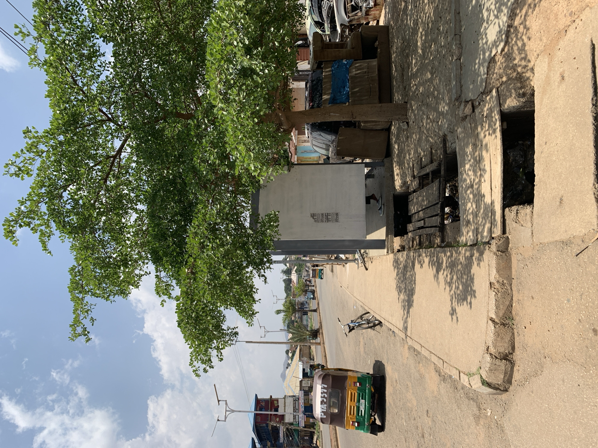 The image shows a street in Rafin Par, Nigeria, during daylight hours. An auto rickshaw is on the right side of the street. A bicycle is lying on the street. A tree is on the left side of the road and extends over the road. Next to the tree are stacks of what seem to be objects. Beyond the auto rickshaw are buildings, power poles, and other vehicles. The sky is partly cloudy.
