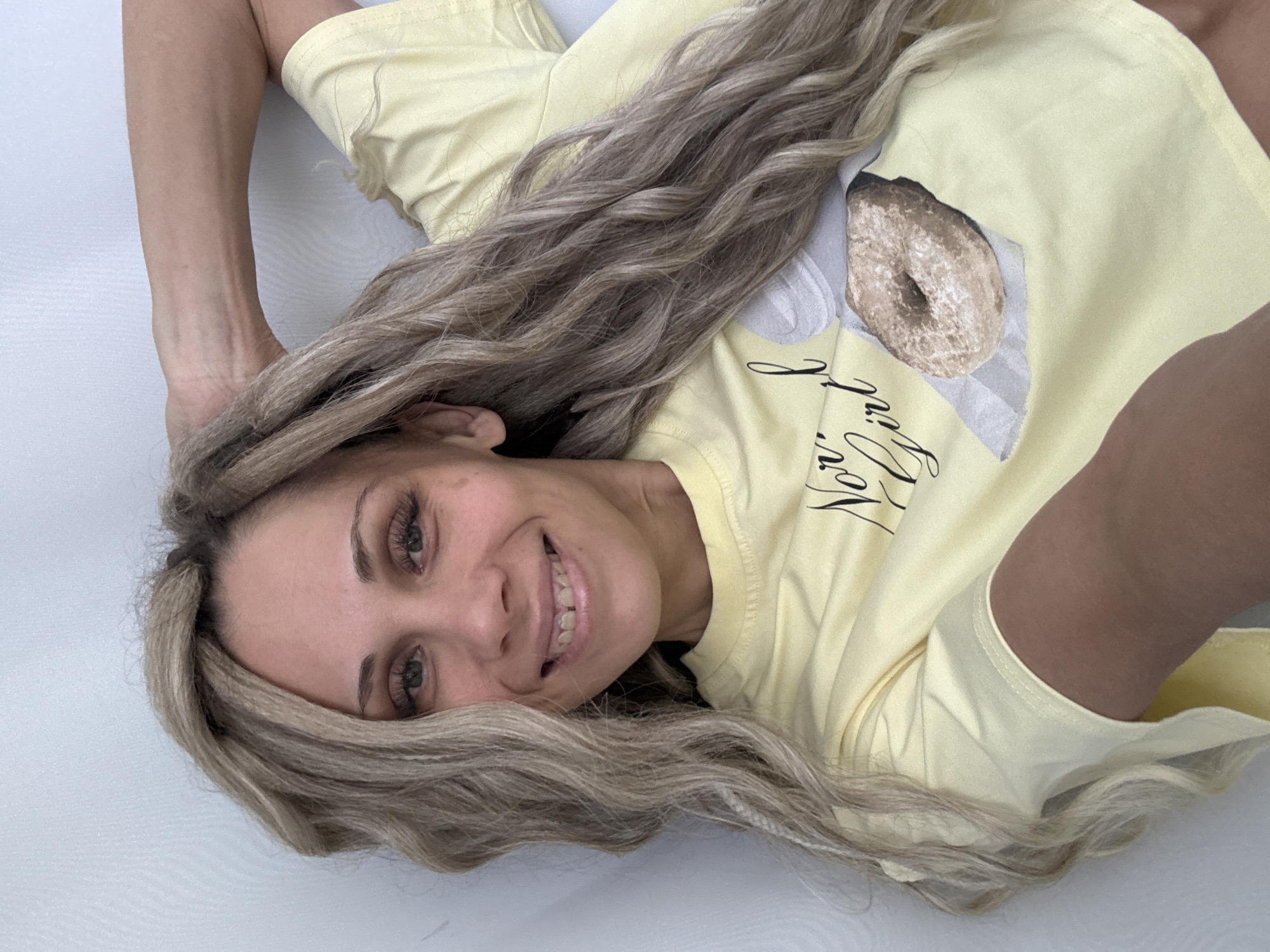 A woman with long, blonde hair is lying on a white surface. She is wearing a yellow t-shirt with a donut graphic. She is smiling at the camera. The location is unknown, and there is no evidence to suggest the image was taken in Rusanovo, Russia.