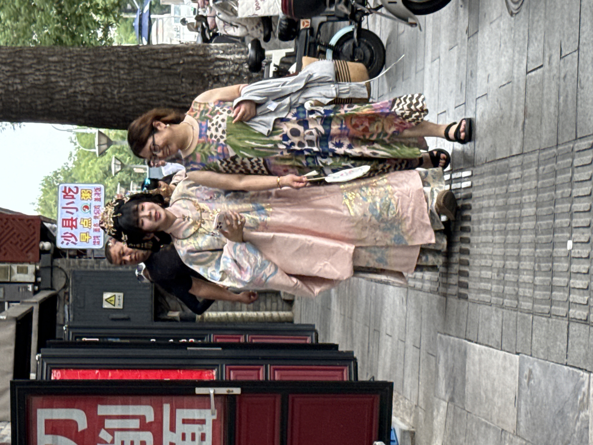 This image captures a street scene in Xicheng District, China, likely during the daytime given the bright lighting. Two women are the primary subjects, dressed in traditional and modern attire, suggesting a cultural event or a themed photoshoot. The woman in the foreground wears a pink traditional-style dress and elaborate headwear, resembling a costume. The woman behind her is in a more contemporary dress with a vibrant floral pattern and a light jacket draped over her shoulders. Both women appear to be posing or interacting with each other.
The setting is an urban environment, characterized by a paved sidewalk, a tree trunk on the left, and shopfronts in the background. Several signs are visible, with some text readable. One sign prominently displays "沙县小吃" (Shaxian Snacks), a popular fast-food chain in China. Other legible text includes "早点粥" (breakfast congee) and "爐饰面多保焖鸡 盖浇饭" (likely indicating various dishes like stewed chicken and rice bowls). The presence of these signs suggests the area is commercial, with food establishments. A scooter or motorcycle is also partially visible in the background, a common sight in Chinese cities. The overall mood seems relaxed and pleasant, with the women smiling and appearing engaged. The weather appears to be clear, with ample sunlight casting shadows.