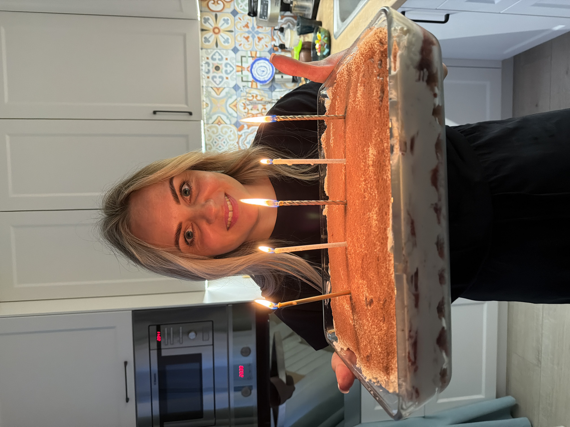 A woman with blonde hair is holding a rectangular glass baking dish with a cake inside. The cake appears to be dusted with a brown powder, likely cocoa, and has layers of white cream and what appears to be red fruit. Six lit candles are inserted into the cake. The woman is smiling at the camera. She is wearing a black top.
Behind her, there are white kitchen cabinets and a colorful tiled backsplash. A microwave oven is visible on the wall below the cabinets. A blue dish is mounted on the backsplash. The floor is a light-colored tile.
There is nothing in the image to suggest this is in Votkinsk, Russia.