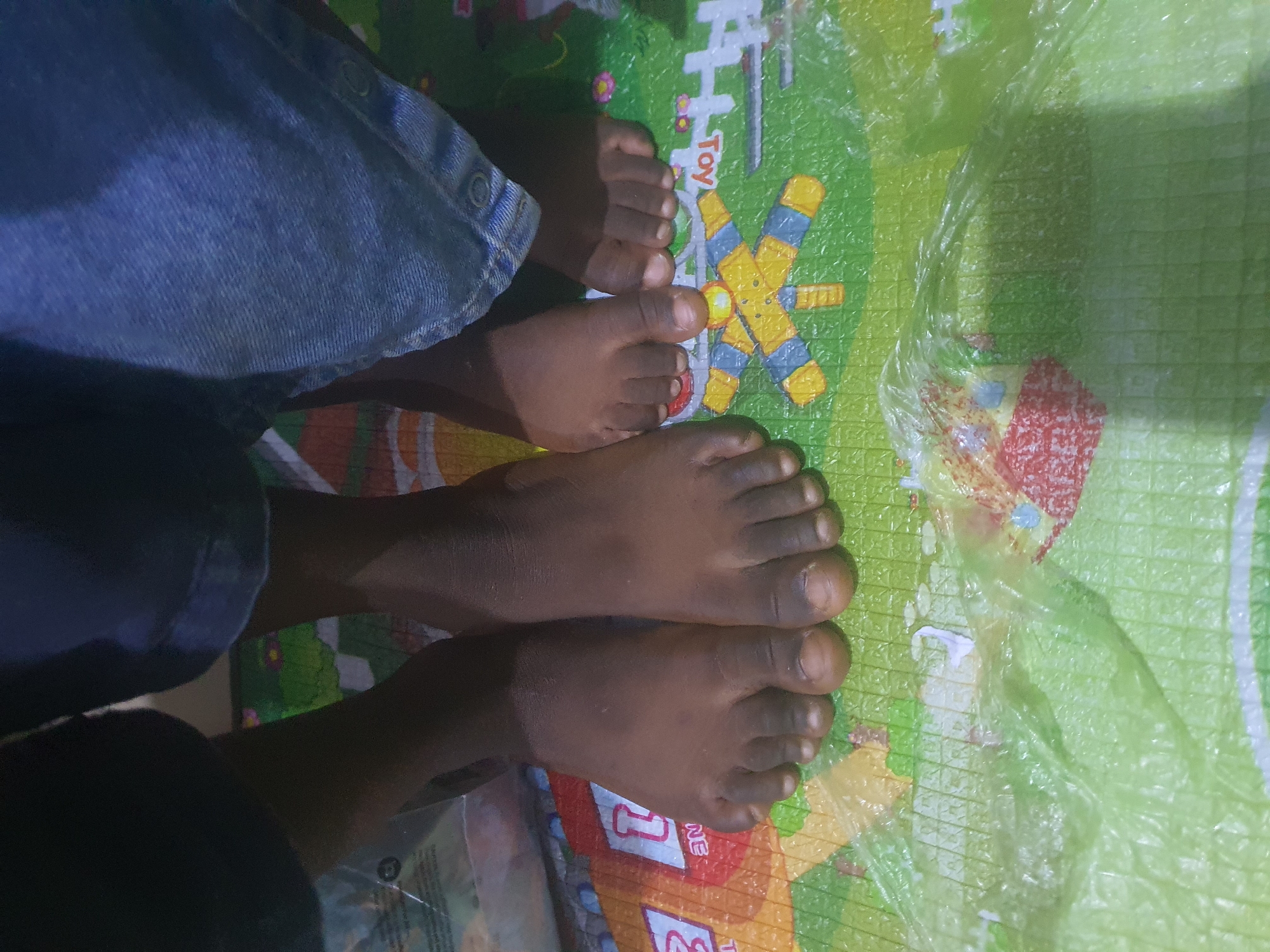 The image shows a close-up view of two pairs of bare feet and the lower legs of two individuals, one pair of feet appears to be larger than the other, suggesting different ages or sizes. The individuals are wearing blue denim pants. They are standing on a colorful play mat with cartoon-like illustrations, possibly a children's educational mat, featuring graphics that include letters, numbers, and simple shapes like a yellow and blue robot-like figure and a white structure that resembles a house. A crinkled, translucent green plastic sheet or bag partially covers the foreground of the mat. The scene is indoors, likely in a residential setting in Jos, Nigeria.