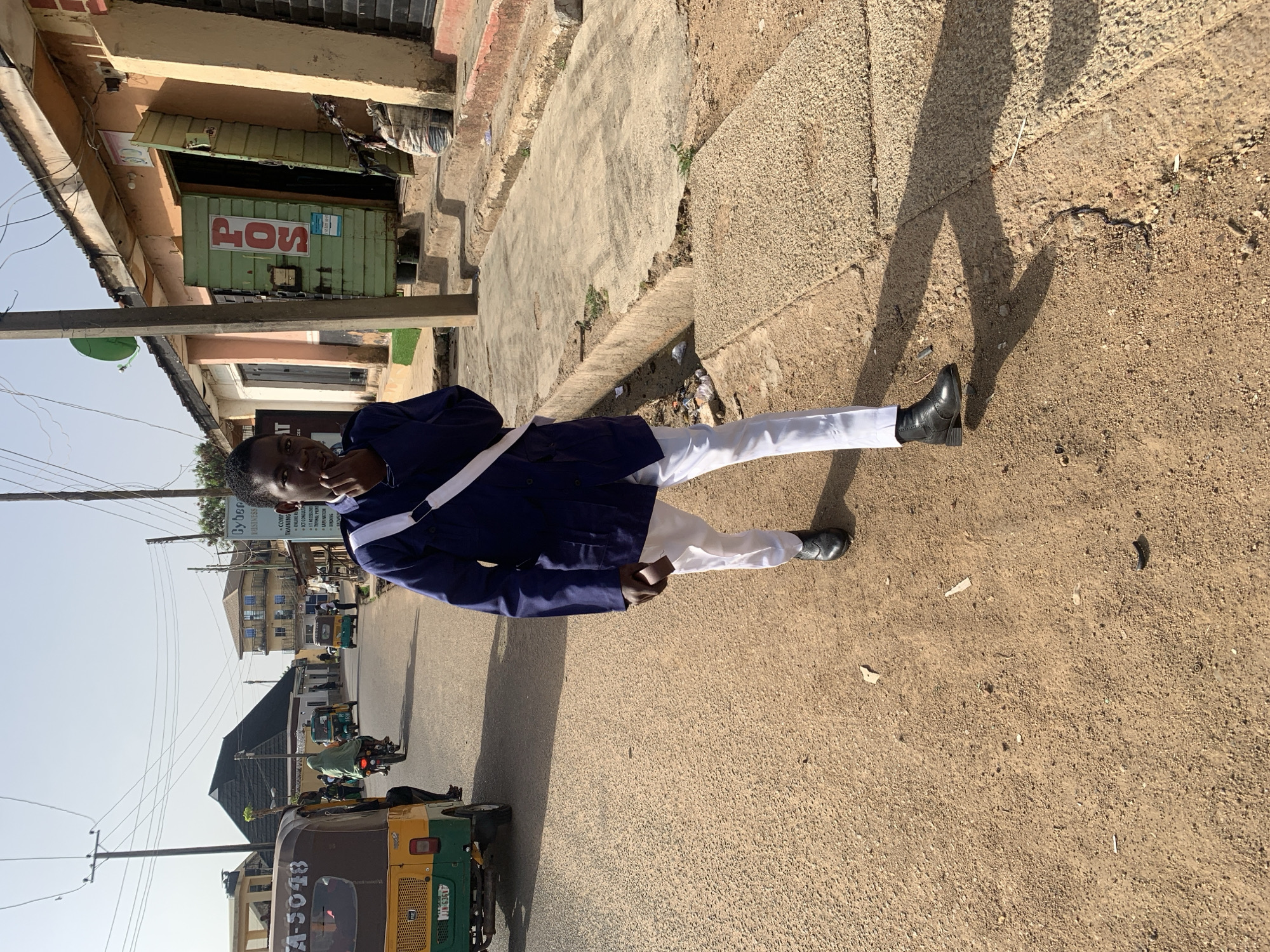 A young man stands on a paved street in Jos, Nigeria, with a building to his left featuring a "POS" (Point of Sale) sign. He wears white pants and a dark blue jacket with a white strap across his chest. He appears to be holding a small brown object in his hand. The street scene includes a yellow and green vehicle with "A-5048" visible on its side. Several other vehicles and people are visible in the distance. Above the vehicle, a sign for "Cyber Business" is visible, listing services like "Computer Training" and "Online Registration." The street is covered with loose dirt and scattered debris.
