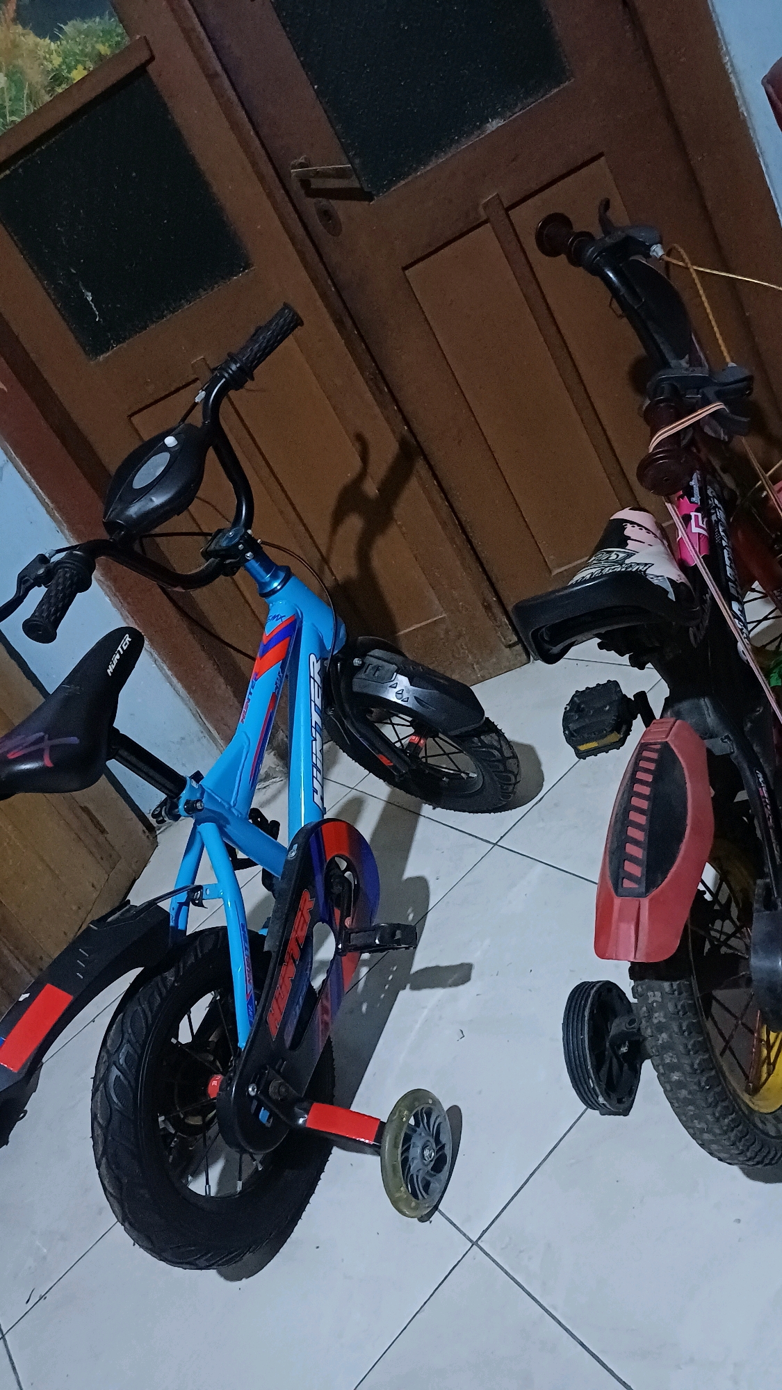 Two children's bicycles are parked indoors on a tiled floor. The bicycle on the left is bright blue with red and black accents, featuring training wheels. Its seat is black and bears the "HUNTER" logo. The bicycle on the right is predominantly black with red accents and yellow rims on its wheels, also equipped with training wheels. Both bicycles appear to be of similar size, suggesting they are intended for young riders. The background consists of brown wooden doors. The lighting suggests an indoor environment, possibly a home or storage area. The image is taken from a slightly elevated angle, looking down at the bicycles. There are no people visible in the image. The city of Wonokerso, Indonesia, is not directly discernible from the image itself but is provided as contextual information for the location.