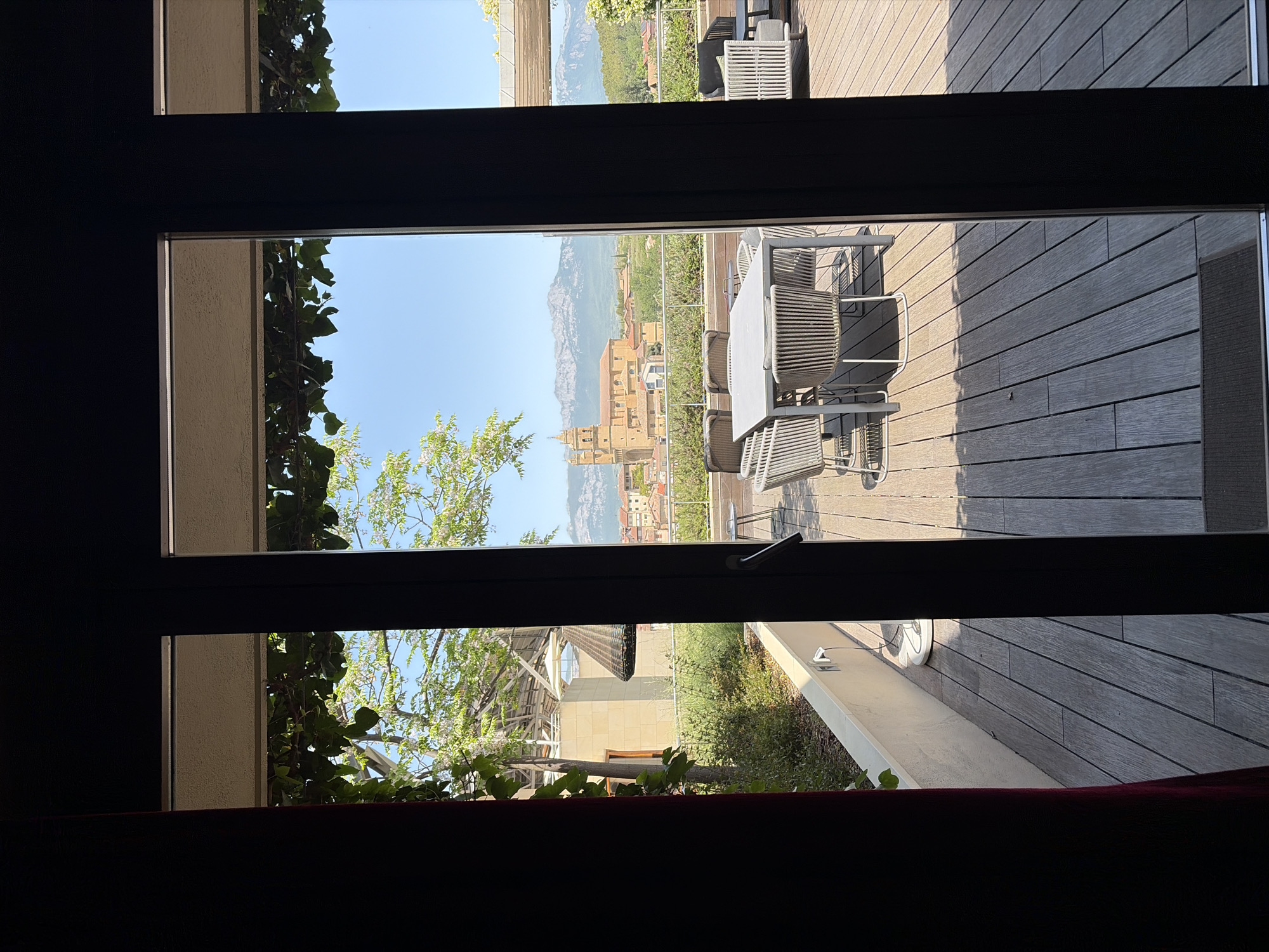 The image depicts a view through a window in Unknown, Unknown. Outside, there's a wooden deck with a table and chairs. In the distance, there is a city with buildings and hills. Greenery is visible in the middle ground. The window frame is dark and obscures parts of the view.