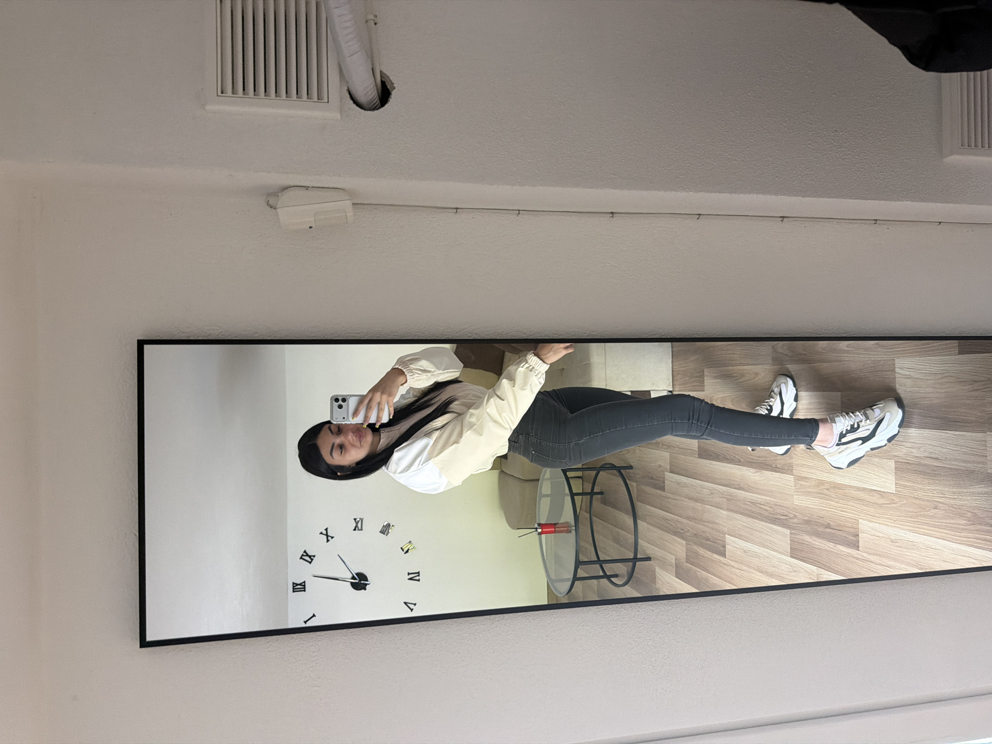 A woman takes a picture of herself in a mirror. The mirror is mounted on the wall and has a clock design on the left side. A small round table is in the reflection near the woman's waist, with a red and silver object on it. The room has a wooden floor, and the walls are white. The specific location in Gabrovo, Bulgaria cannot be determined from the image.