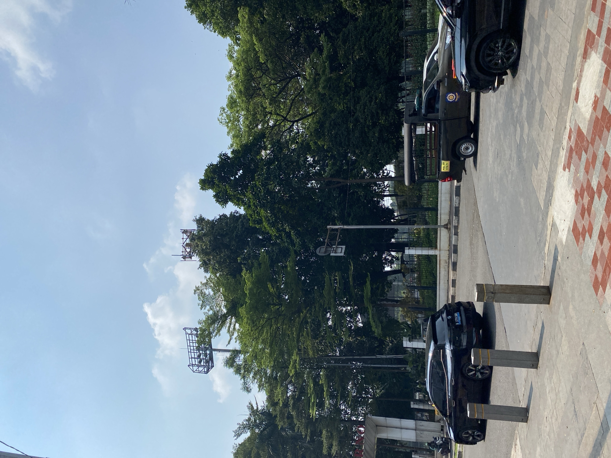 In the city of South Jakarta, Indonesia, a street scene unfolds under a partly cloudy sky. A black SUV, a BMW iX, is parked on the right side of the image, its rear partially visible. To its left, a dark green utility vehicle, likely a police or public order vehicle, is seen from the side. It has an open cargo bed with a frame structure and a label that reads "POL PP 515-71". A white car is partially visible behind the utility vehicle. In the foreground, a sidewalk with patterned tiles leads towards the street, with three concrete bollards in a row. The background is dominated by a dense collection of green trees and two tall structures with metal frameworks, possibly lighting towers for a sports field. A partially visible entrance gate with a white facade is on the left side of the image. A person on a motorcycle is also seen on the far left.
