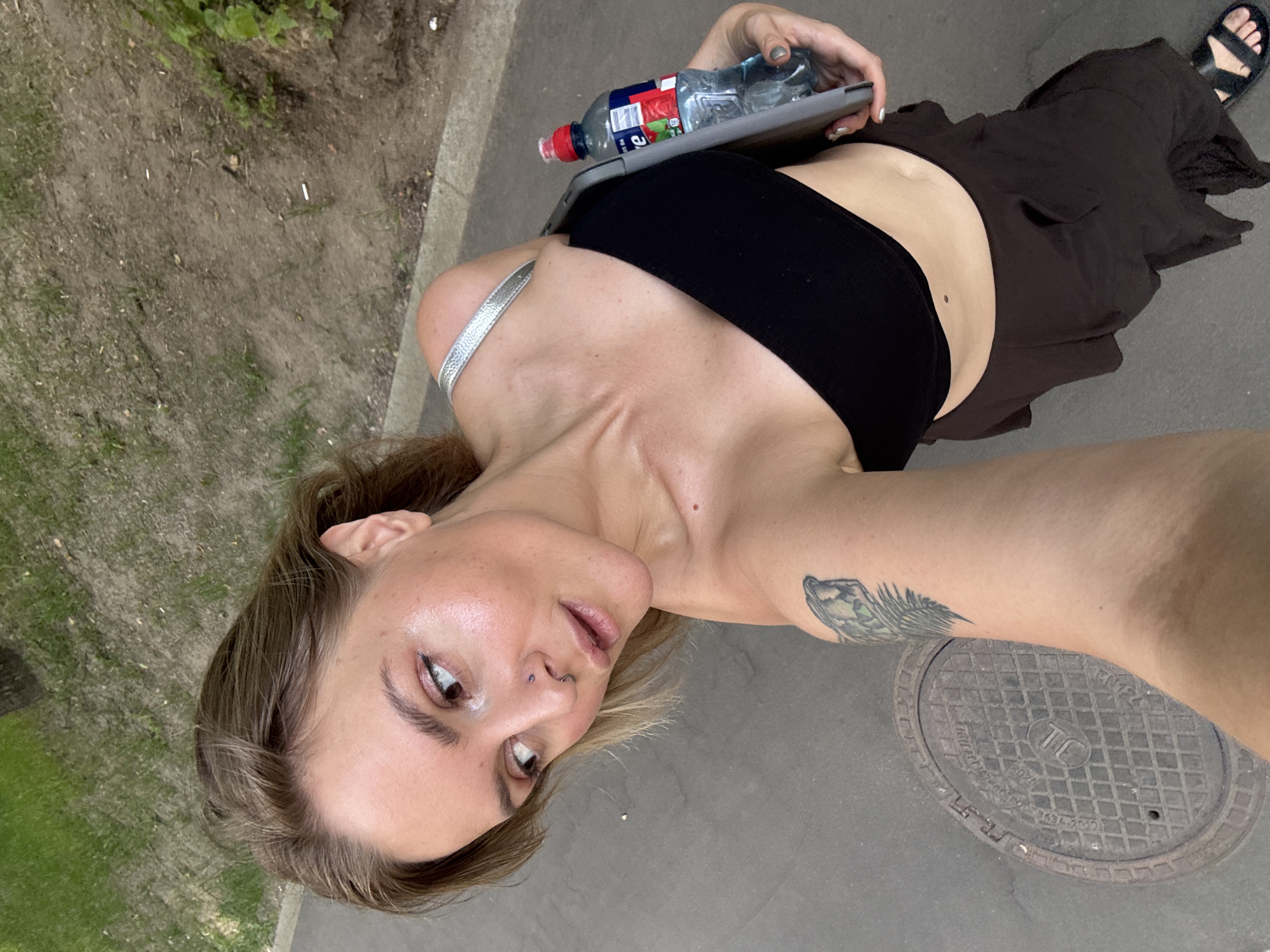 A woman with blonde hair is taking a selfie outdoors. She is wearing a black strapless top and a brown skirt. She has a tattoo on her left arm and is holding a tablet and a bottle of water. The background shows a paved area with grass and a manhole cover. The image was taken on a sunny day.