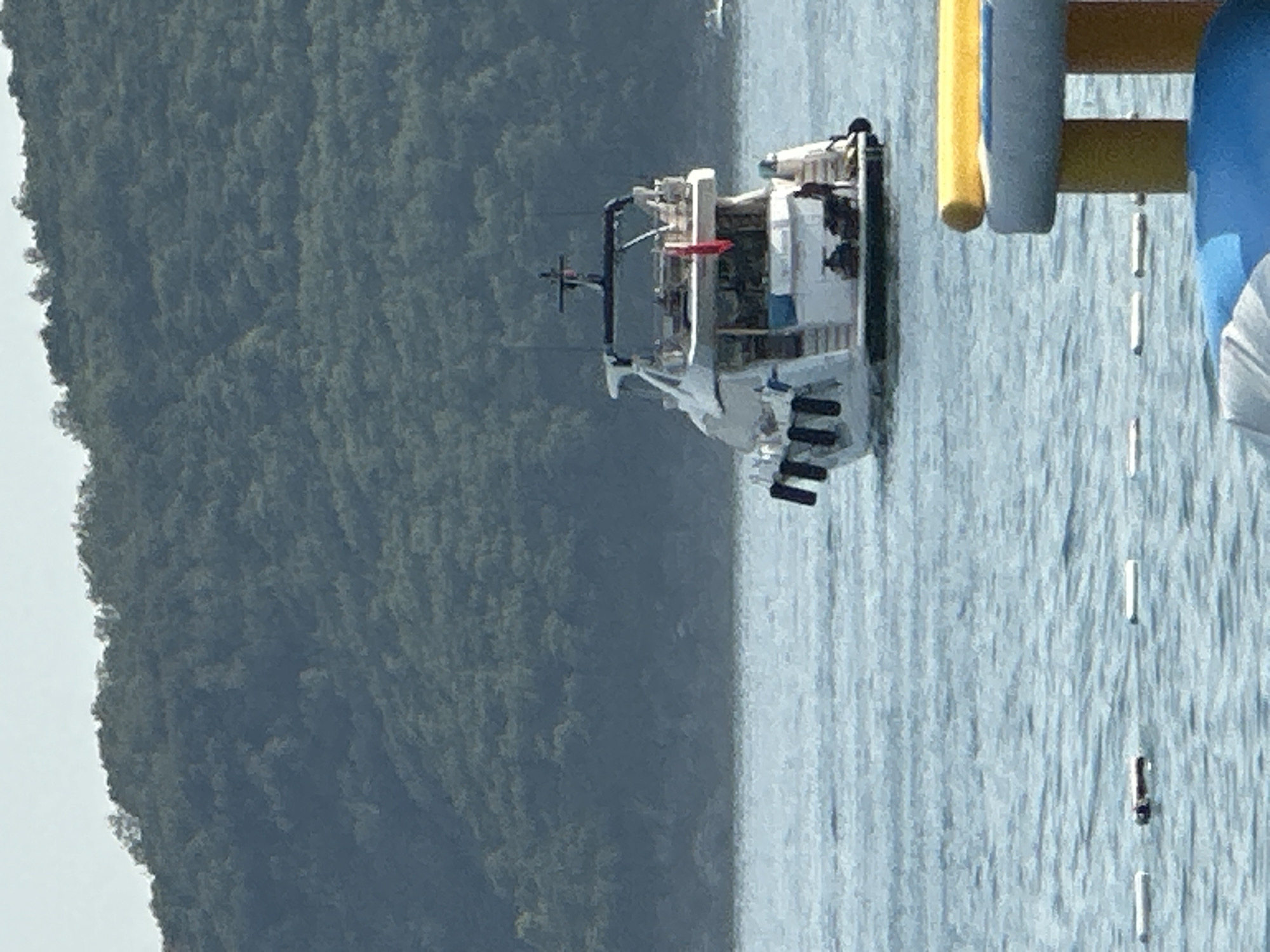 In Boreti, Montenegro, a white yacht is moored in the water, with a dense, green, tree-covered hill rising behind it. The water is calm and blue, with a few white buoys floating on the surface. In the foreground, a portion of a yellow and blue inflatable water toy is visible. A single person is standing on the stern of the yacht. The sky is a pale, hazy blue.