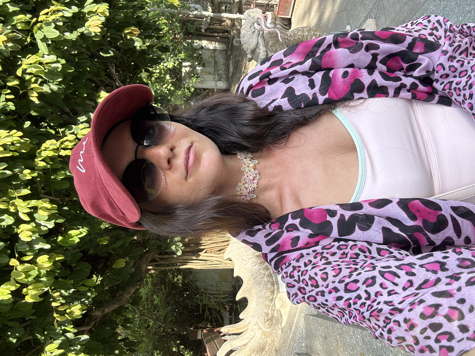A woman is taking a selfie in an outdoor setting. She is wearing a maroon baseball cap with white lettering, sunglasses, a pink and purple leopard print scarf over a light pink tank top, and a delicate multi-colored beaded choker necklace. Behind her, large green leaves from a tree are visible, as well as a stone sculpture of a dragon. The woman appears to be in the courtyard of a temple or historic site. The lighting suggests it is daytime. The background shows some stone structures and vegetation, characteristic of a tropical environment. Based on the architectural style and the presence of dragon motifs, this scene is likely located at the Long Son Pagoda or a similar Buddhist temple in Nha Trang, Vietnam.