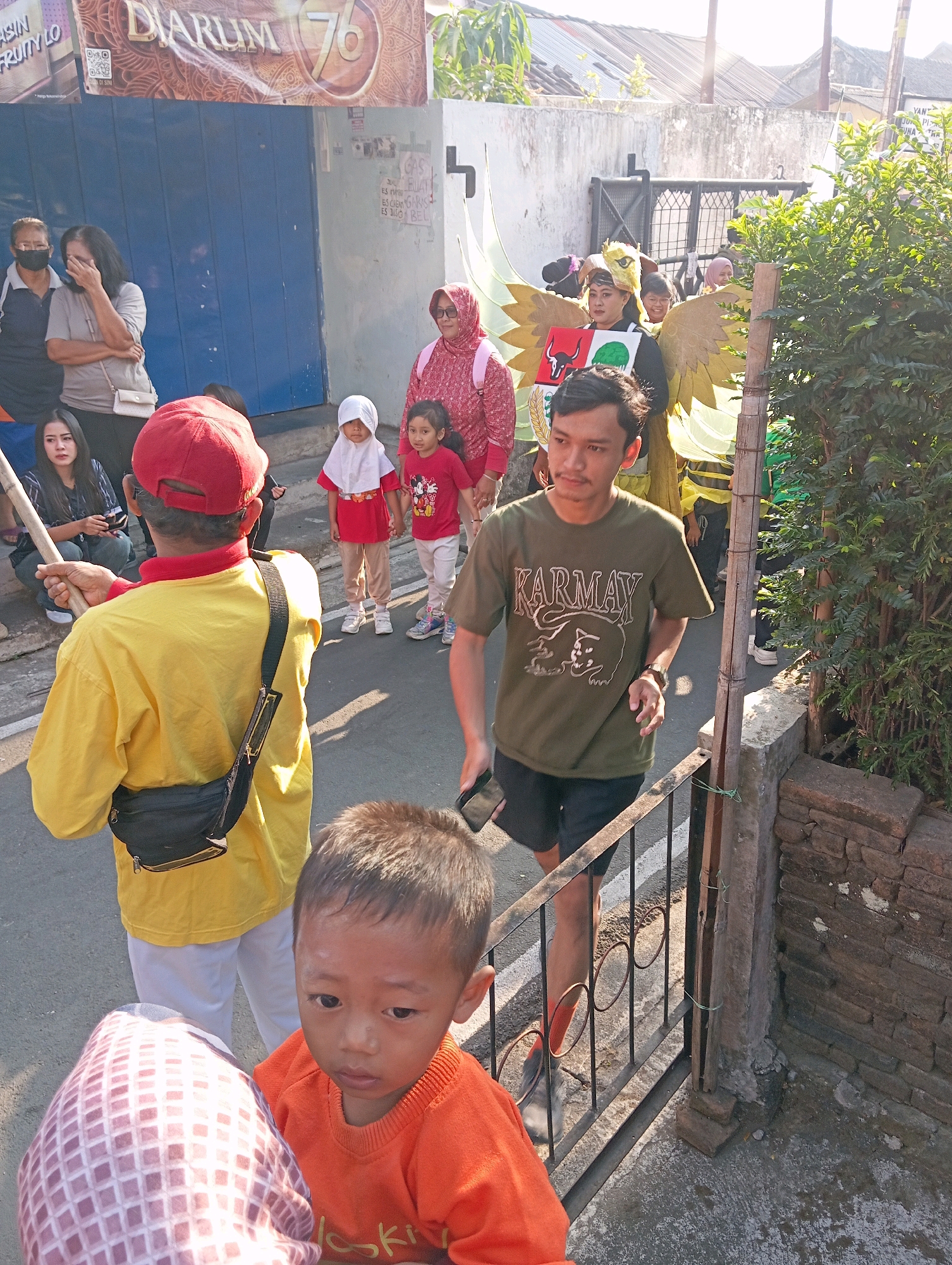 A group of people are gathered on a paved street in what appears to be the city of Magelang, Indonesia. In the foreground, a young child in an orange shirt looks directly at the camera. Behind the child, an adult wearing a yellow shirt and a red cap is holding a wooden pole. Further back, a group of people are observing a procession. One individual is dressed in a costume with large yellow wings, resembling a bird or mythical creature, and holds a shield. Other individuals in the background are dressed in casual clothing. The environment includes a blue building with signage, a stone wall, and lush green foliage. The scene suggests a street event or parade is taking place.