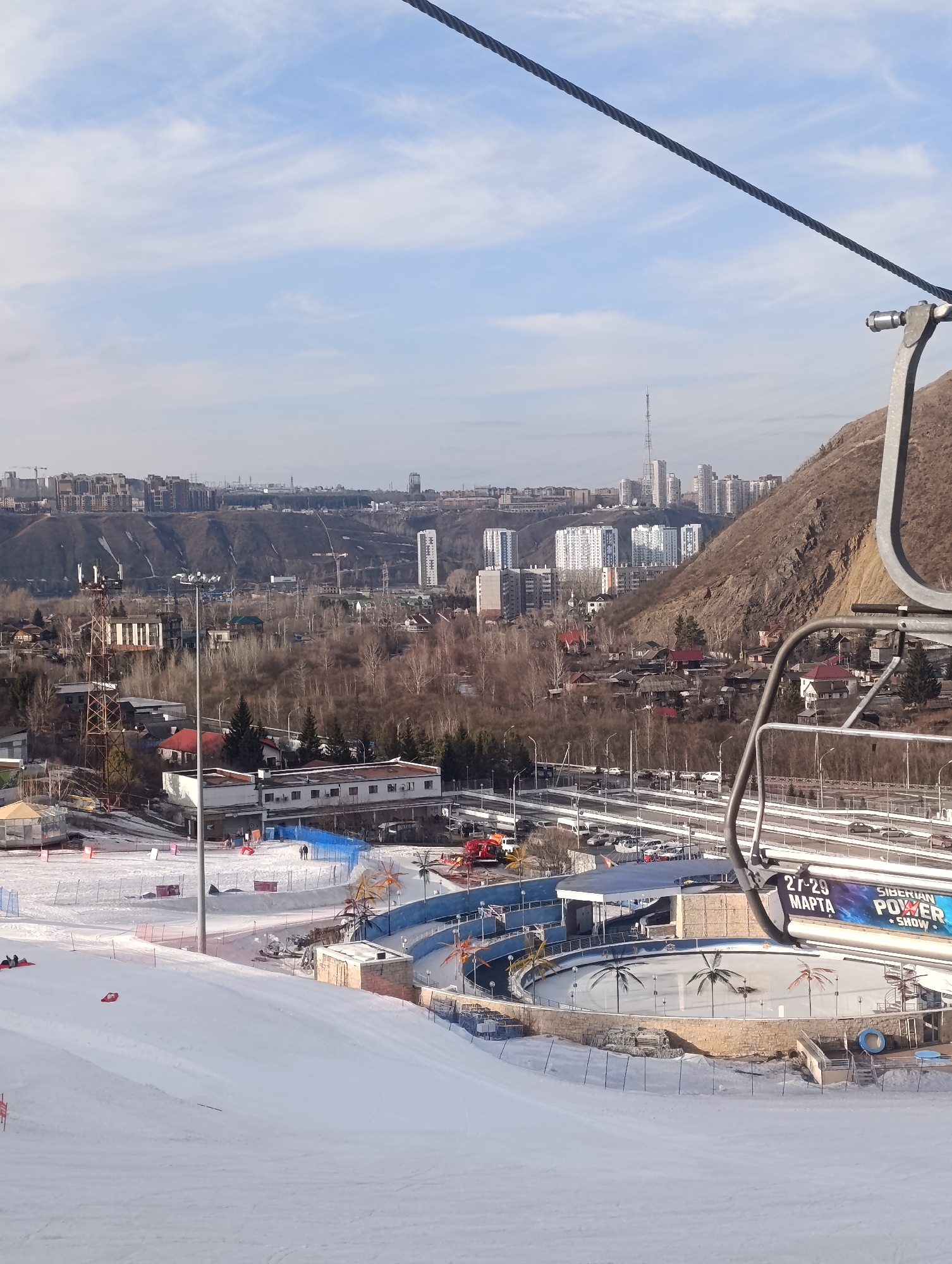 The image depicts a winter panoramic view from a ski lift at the Bobrovy Log Funpark in Krasnoyarsk, Russia. The foreground is dominated by a wide, snow-covered ski slope, featuring two individuals lying on red sleds/tubes near the left edge and two small figures walking uphill further to the right. Orange safety netting and reddish markers are visible on the snow.
In the midground, at the base of the slope, an oval-shaped outdoor rink is present, its surface decorated with painted palm trees. Adjacent to this, a building displays a large blue banner with text "27-29 MAPTA" and "SIBERIAN POWER SHOW." A multi-lane highway with active vehicle traffic and several parked cars runs horizontally across the scene. Various buildings, including a long, white structure and smaller houses with red roofs, are interspersed among leafless trees and a prominent metal communication tower.
The background reveals the extensive cityscape of Krasnoyarsk, stretching across hilly terrain. Numerous multi-story residential and commercial buildings are visible, with a tall, slender TV tower prominently rising above the distant skyline. The hillsides are a mix of bare, brown earth and sparse dry vegetation. The sky is partly cloudy with patches of clear blue.