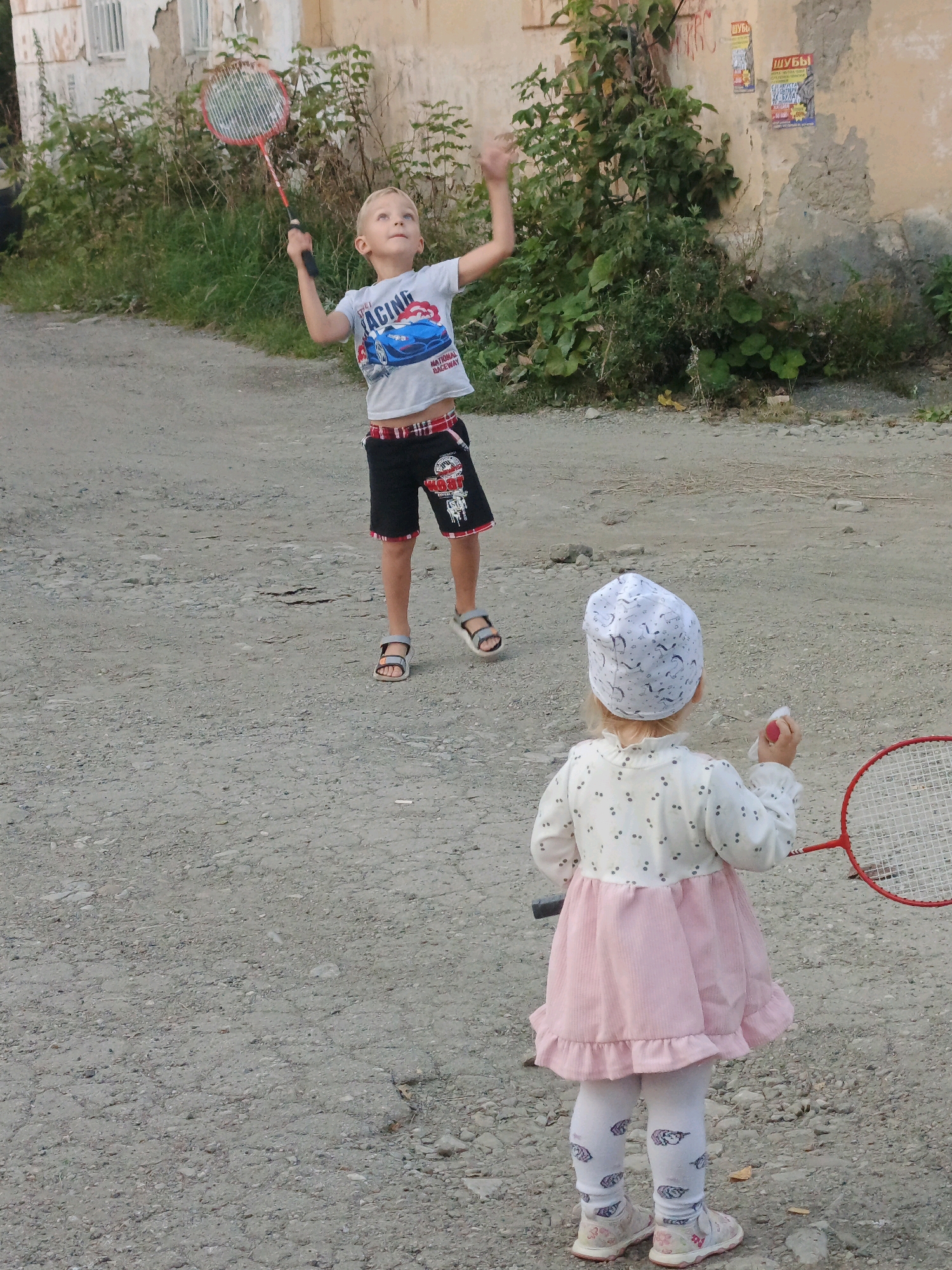 Two children are playing badminton outdoors. A boy, with blonde hair, stands facing the camera, wearing a white t-shirt with a blue car graphic and black shorts. He is mid-swing with a red badminton racket. A younger child, facing away from the camera, wears a white patterned headscarf, a white and pink dress, and white patterned leggings. They are also holding a red badminton racket. They are on a gravel path in front of a building with peeling paint and overgrown vegetation. Two posters are visible on the building wall. This scene is set in Zlatoust, Russia.