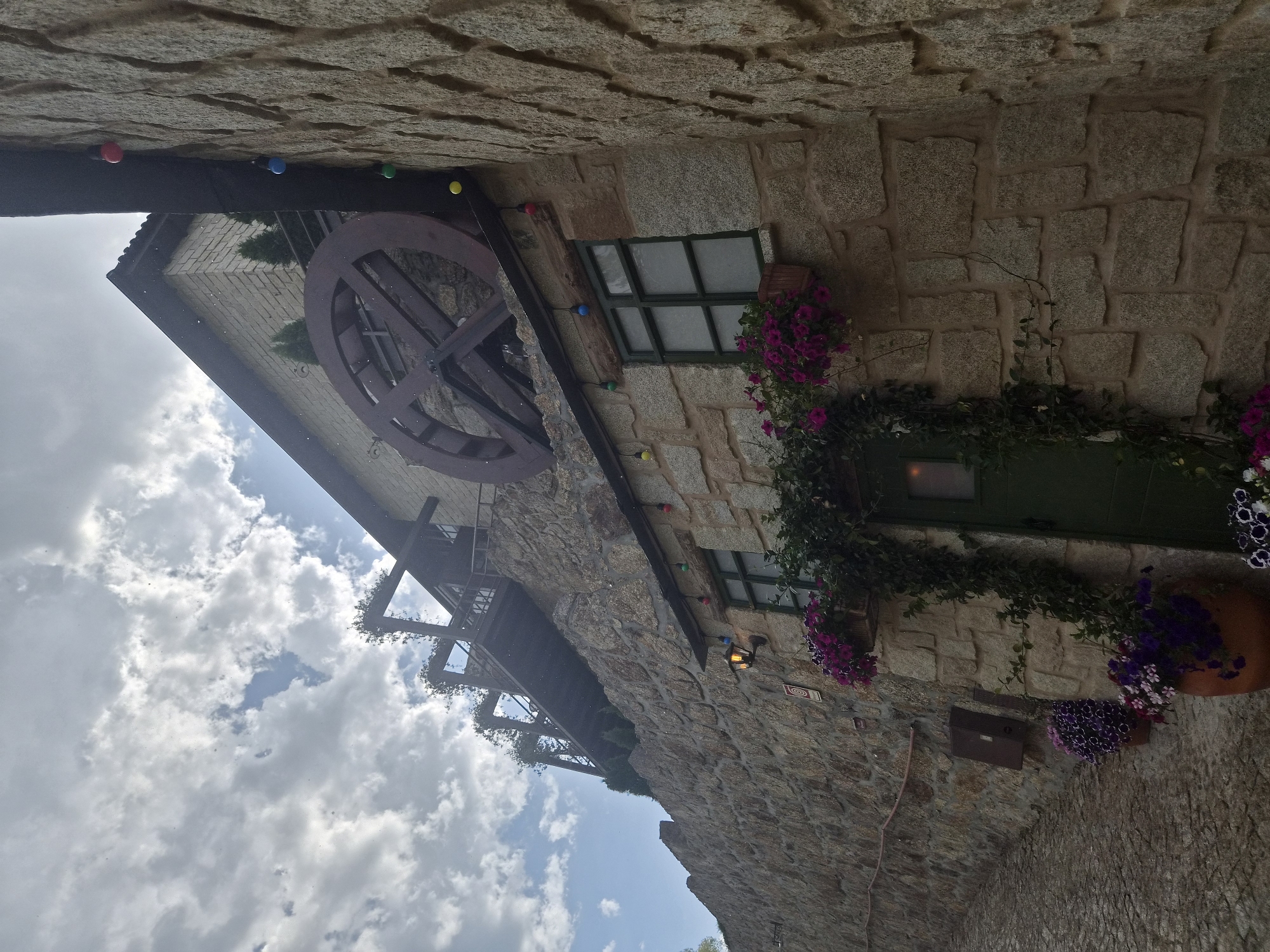 The image depicts a view looking up at a stone building with a large wooden water wheel mounted on its upper section. The building is situated on a steep incline, with a cobblestone path curving to the left in the foreground. String lights with colored bulbs are visible along the building's facade and on a dark wooden beam above a window. Several potted plants with purple and pink flowers are placed near windows and the entrance door, which is painted green. Above the main building, a wooden walkway or balcony structure is visible, partially covered in vegetation. The sky is cloudy, with patches of blue sky showing through. The building appears to be in the historic district of Seia, Portugal, likely a renovated mill or a building designed in a similar rustic style. No people are visible in the image.