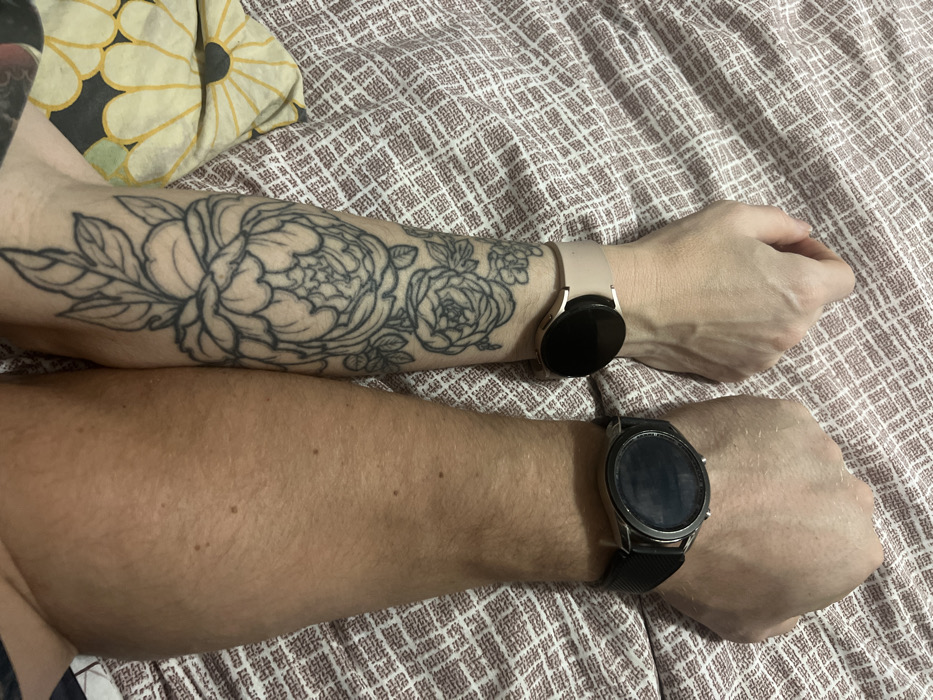 Two forearms and hands are shown resting side-by-side on a textured, light brown or pinkish-hued patterned fabric, likely a bedspread or blanket. In the upper left corner, a yellow and white floral patterned fabric is partially visible, suggesting a pillow or another textile.
The upper arm, which is lighter-skinned, features a prominent black line-work tattoo of large floral designs, including peonies and roses with leaves, extending along the forearm. This arm wears a light pink or beige smartwatch with a circular black screen and a matching band. The hand is relaxed with fingers slightly curled.
The lower arm is darker-skinned and has visible arm hair. It is wearing a dark smartwatch with a metallic silver bezel and a black band. The watch face is dark and appears to be off or in standby mode. This hand is also relaxed, forming a loose fist.
The image captures the forearms and wrists of two individuals, possibly a couple, in an indoor setting, likely a bedroom. There is no discernible activity beyond the arms resting together. No text is visible in the image.