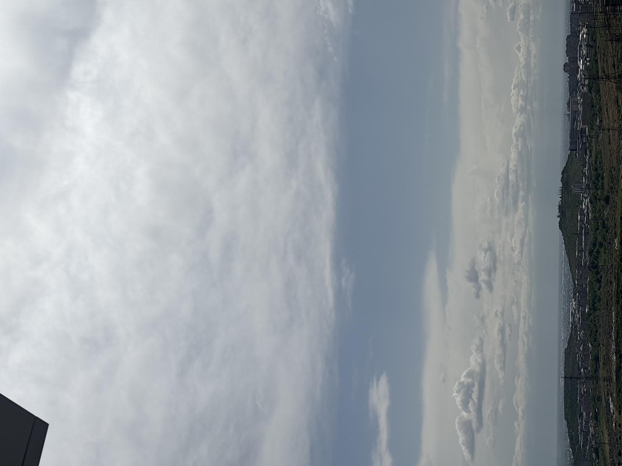 The image shows a sky with varying cloud cover, transitioning from dense, white clouds on the left to a clearer, blue section in the middle, and more layered clouds near the right edge. The right side of the image shows a cityscape with buildings and green areas, likely a park or undeveloped land. The perspective suggests an elevated vantage point. Based on the visual characteristics of the buildings and landscape, the city is likely Yerevan, Armenia. Power lines are visible in the right foreground, adding to the urban scene. A dark structure is visible in the lower left corner.