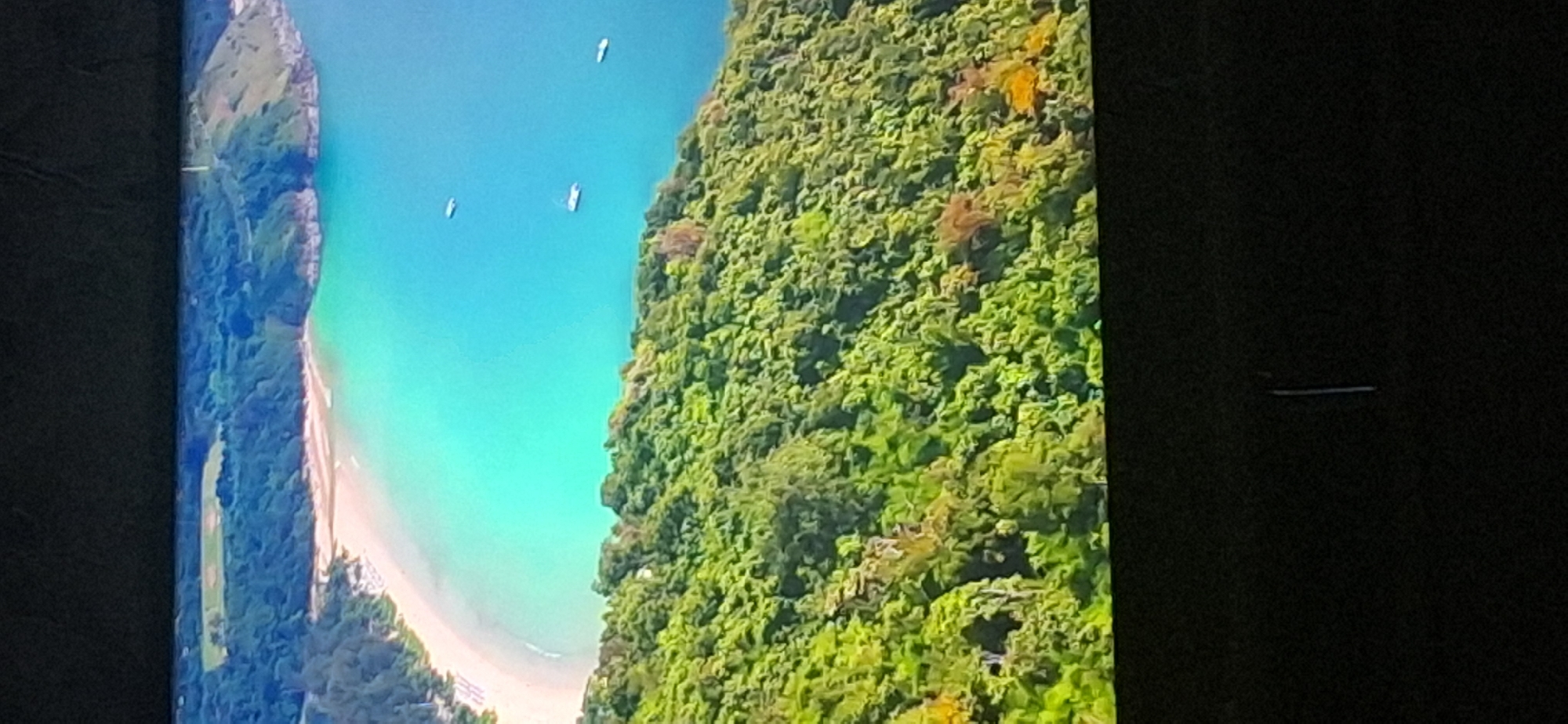 This is a photograph of a tropical coastline from an aerial perspective. A curved, sandy beach meets a calm, turquoise body of water. Lush green foliage covers the rolling hills that extend from the coastline. Several small boats are visible on the water. The image is taken from a high vantage point, looking down and across the landscape. There is no information available about the city of Bila Tserkva, Ukraine, in relation to this image.