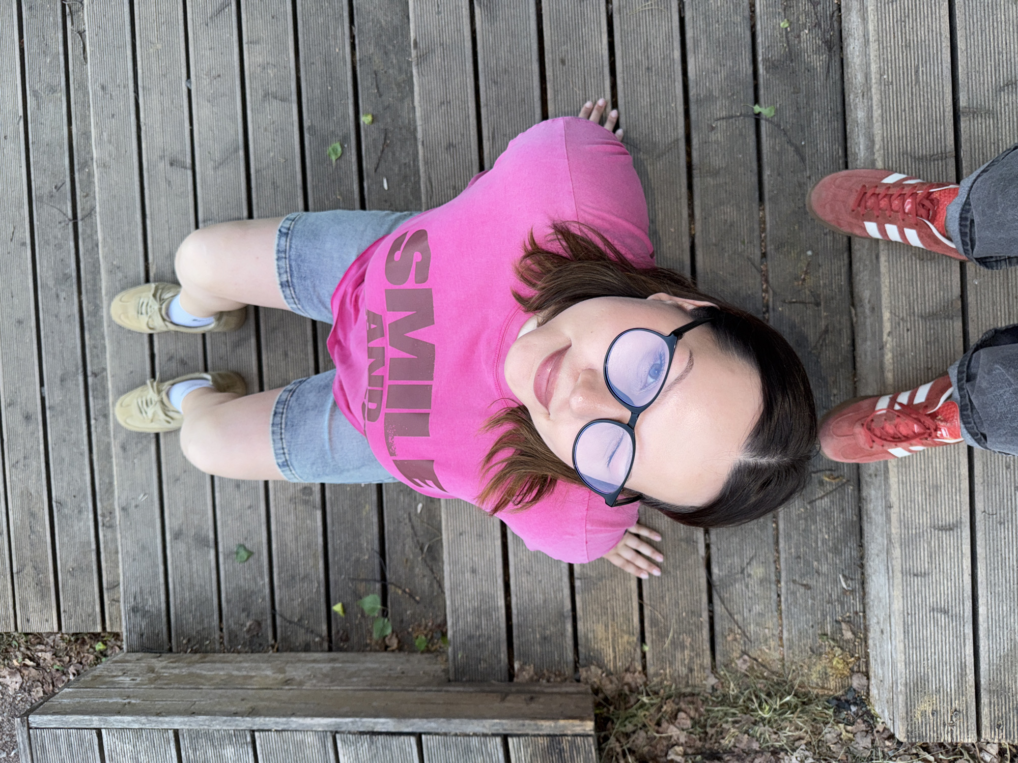 A top-down, eye-level shot depicts two people sitting on a wooden plank structure in what appears to be a park-like setting. One person is in the center of the frame, wearing a pink t-shirt with a word partially visible, denim shorts, tan sneakers, and black framed eyeglasses. The second person is partially visible at the top right of the frame, wearing red sneakers with white stripes and gray jeans. The background shows the wooden planks and a glimpse of foliage along the perimeter of the wooden area. Based on visual context and available information, the location is suspected to be in the city of Chaikovsky, Russia.