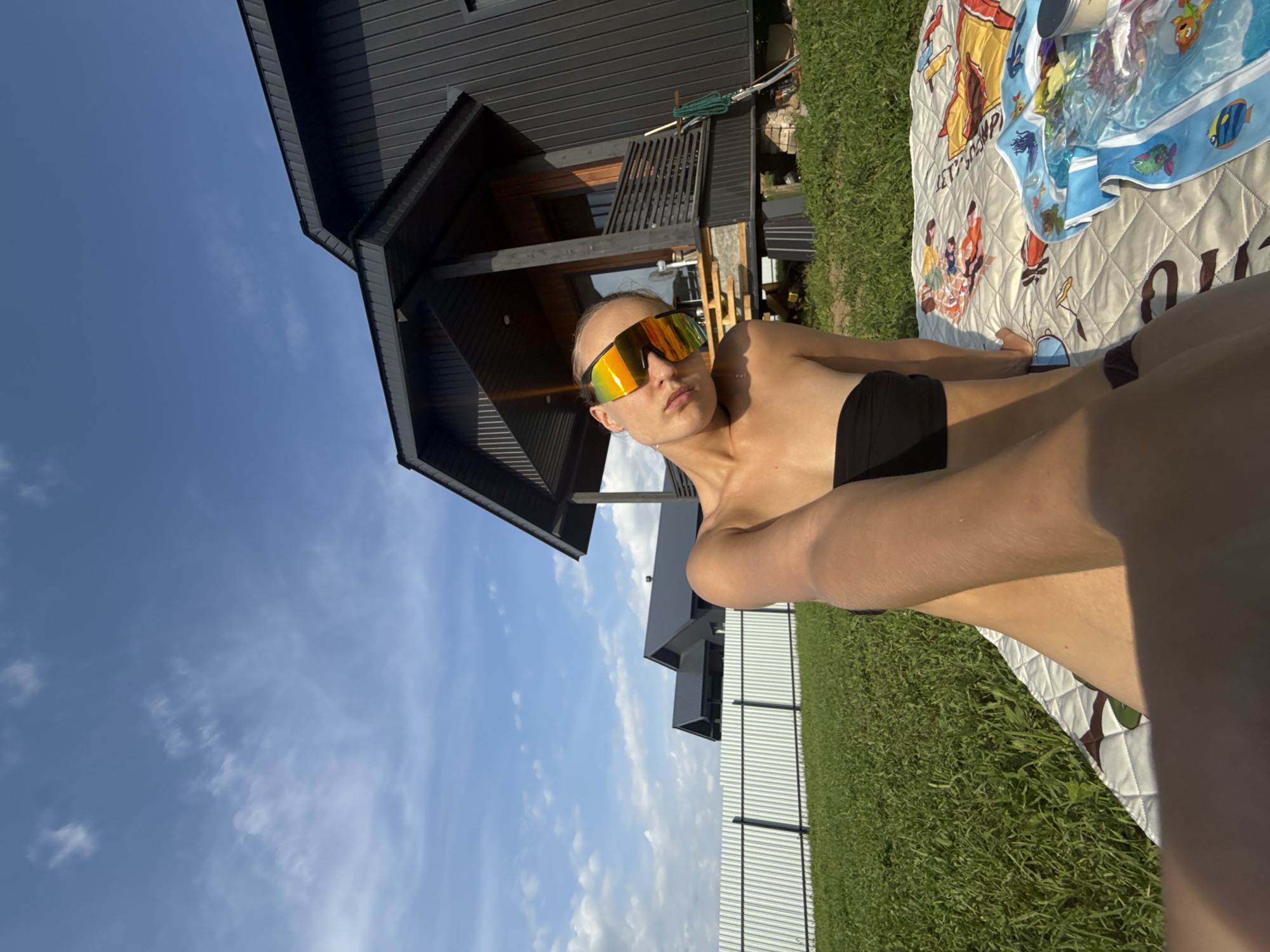 In Подшивалово, Russia, on a sunny day, a young woman with blonde hair and reflective sunglasses is sitting on a patterned blanket in a grassy yard. She is wearing a black bandeau bikini top. Behind her is a modern, dark-colored house with a wooden porch and a white fence. The sky is blue with scattered white clouds. The woman is holding her arm up, as if taking a selfie.