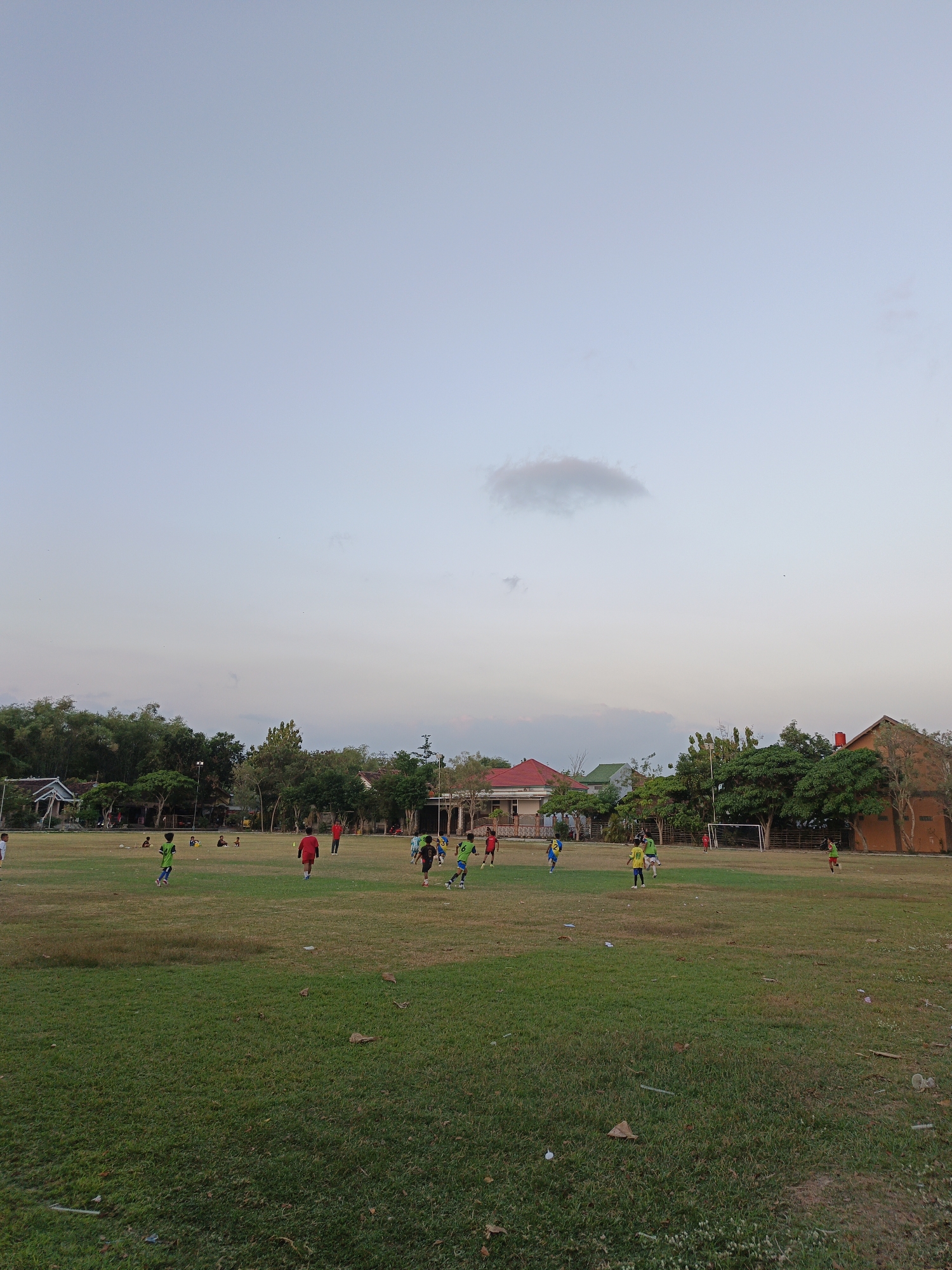 The image depicts a wide-angle view of a grassy field where several young individuals are engaged in a game of soccer. The setting appears to be a rural or semi-rural area, possibly in Planggu, Indonesia, given the architectural style of the buildings visible in the background. The sky is overcast with a light, muted palette of blues and grays, with a faint, wispy cloud formation in the upper portion of the frame.
In the mid-ground, a group of approximately ten to twelve children, mostly boys, are scattered across the field, actively participating in the soccer game. They are wearing brightly colored jerseys, predominantly green and yellow, with some wearing red. Some are running, others are kicking or chasing a soccer ball, indicating dynamic movement and interaction within the game. A soccer goal is visible on the right side of the field, suggesting a designated playing area.
The foreground is dominated by a lush green lawn, with some patches of dry, yellowish grass and scattered fallen leaves, indicating a natural, unmanicured environment. In the background, a line of trees forms a backdrop to the field, with some houses and buildings interspersed among them. The buildings exhibit a typical Southeast Asian style with pitched roofs. A larger, reddish-brown brick structure is prominently featured on the right side of the background. The overall atmosphere suggests a casual, recreational activity taking place in the late afternoon or early evening, with the soft lighting indicative of a fading sun. The scene is devoid of any adult supervision explicitly visible.