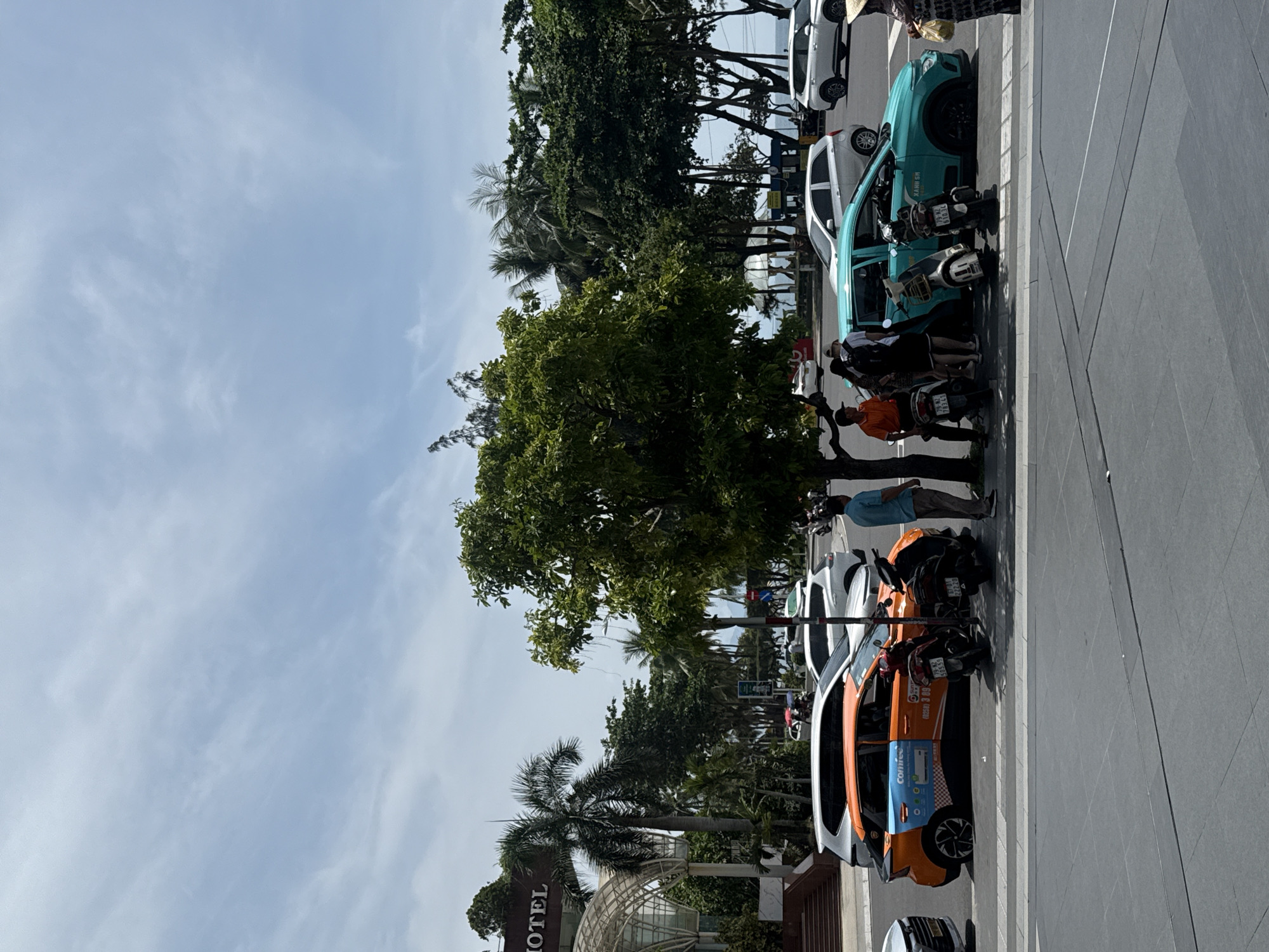 This image captures a street scene in Nha Trang Ward, Vietnam, under a partly cloudy sky. In the foreground, a gray sidewalk is visible. Beyond the sidewalk, a street is populated with various vehicles, including cars and motorcycles.
Dominating the midground is a large tree with dense green foliage, casting shadows on the street. To the left of the tree, an orange car with a blue and white advertisement is parked. Next to it, two motorcycles are parked, one black and one red.
Several people are interacting in the area around the vehicles. Some are standing near the orange car and the motorcycles. To the right of the tree, a turquoise car with Vietnamese writing on its side is parked, with people standing near it. Another person in a straw hat is visible on the far right of the frame.
In the background, there are more trees, including palm trees, and glimpses of buildings and the ocean. A sign with the word "HOTEL" is visible on a building on the far left. The overall scene suggests a busy urban environment with a tropical atmosphere.