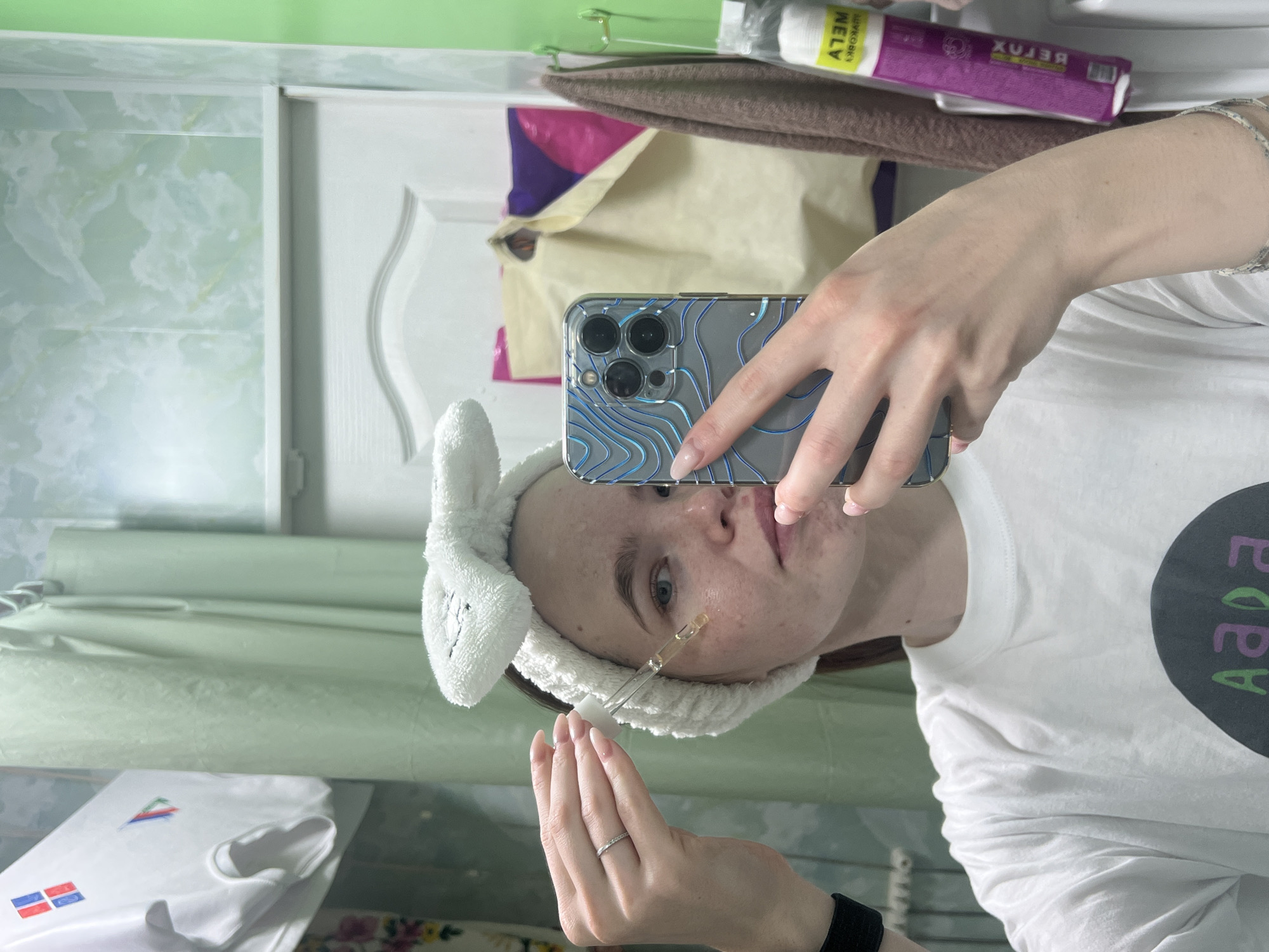 A person is taking a selfie in a bathroom. The bathroom has green and white marble-like walls, and a white cabinet. The person is wearing a white t-shirt with black and green text, and a white bunny-eared headband. They are applying serum to their face with a dropper. The phone case is clear with a blue topographic design. The location is Chaikovsky, Russia.