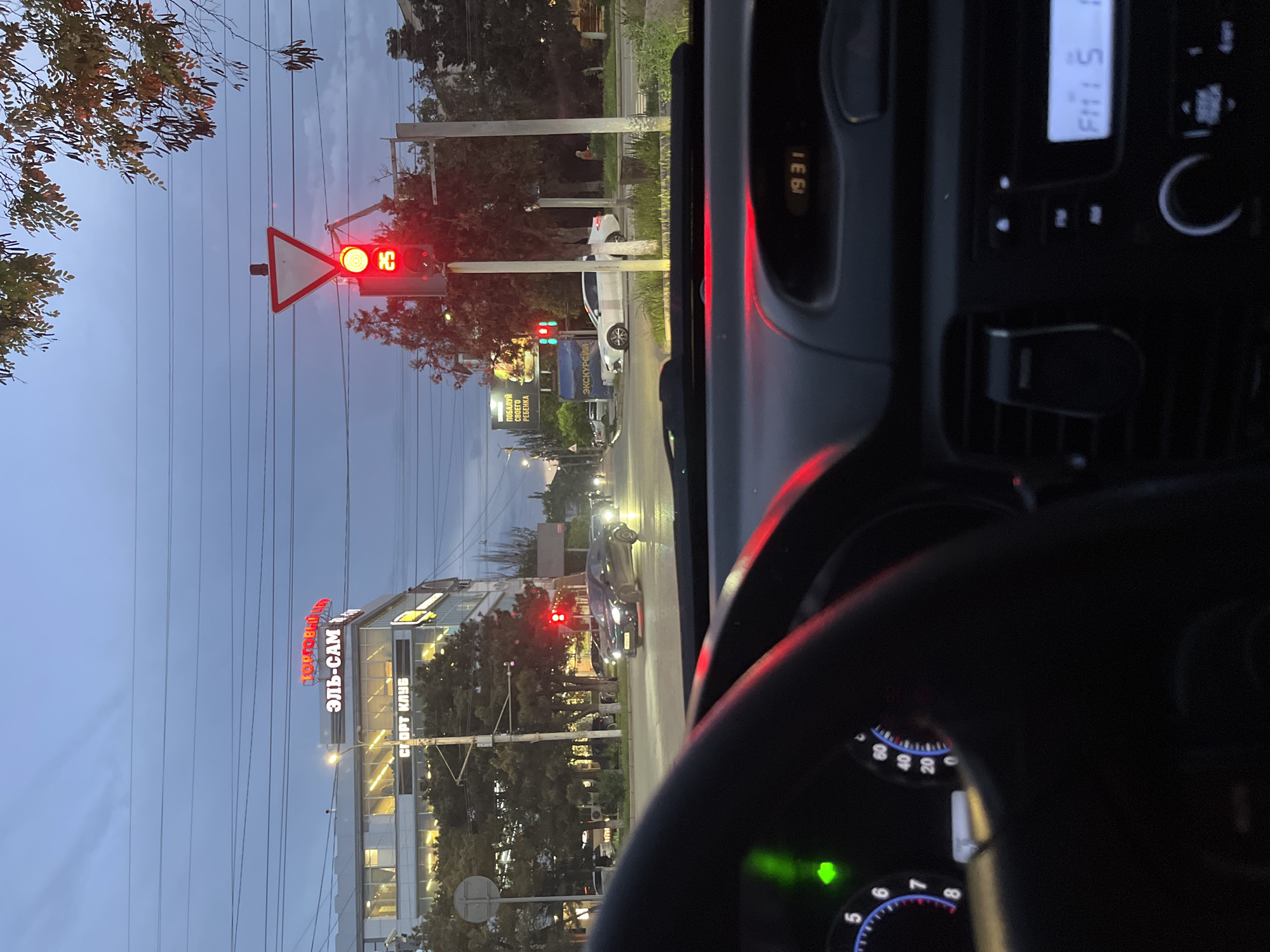 The image shows a street scene in the city of Kaspiysk, Russia, taken from inside a vehicle. The time displayed on the car's dashboard is 19:31. The radio displays "FM: 5". The car is stopped at a traffic light, which is currently red. The yield sign is visible near the traffic light. In the background, a building is visible with a red sign that reads "ТОРГОВЫЙ ЭЛЬ-САМ СПОРТ КЛУБ", indicating a commercial center. A billboard is visible that reads "ПОБАЛУЙ СВОЕГО РЕБЕНКА ЭКСКУРСИИ". Other vehicles are on the road. The environment appears to be urban. Overhead power lines are visible against the sky.