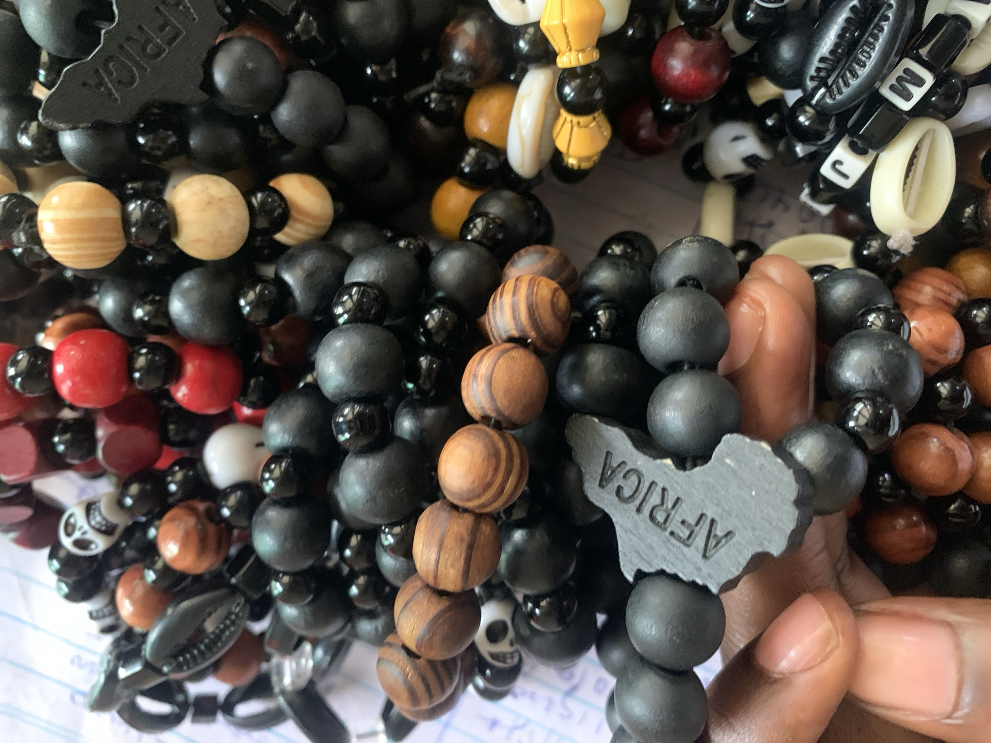 The image shows a collection of beaded bracelets being held in a person's hand. The bracelets are made of various materials and colors, including black, brown, red, white, and gold. Some bracelets feature cowrie shells and lettered beads, while one bracelet has a charm in the shape of the African continent with the word "AFRICA" inscribed. The setting suggests an informal, possibly commercial context, given the variety of bracelets available. The presence of the "AFRICA" charm potentially indicates a connection to African heritage or culture. Although it has been reported that the location is in the city of Kabong, Nigeria, there are no details in the image that confirm it.