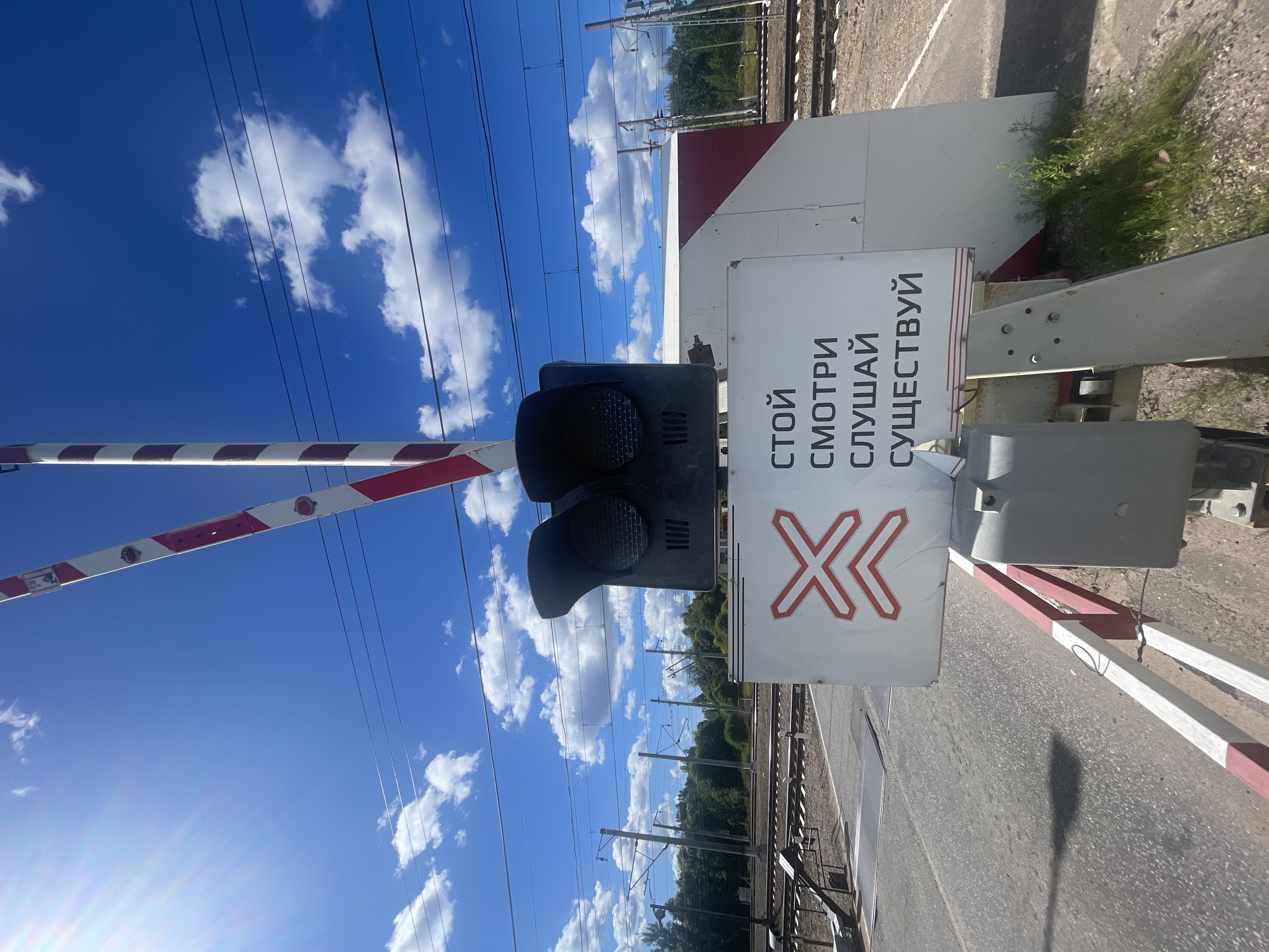 The image depicts a railroad crossing with a barrier arm that is red and white striped and raised. The barrier arm is angled upwards, suggesting it is either in the process of opening or closed. Above the arm, two black signal lights are visible. In front of the barrier, a white sign with red markings is displayed. The sign features a red cross symbol with chevrons pointing inwards on the left side. To the right of the symbol, black text is printed in Russian, which translates to "STOP. LOOK. LISTEN. EXIST." The sign is positioned on a concrete structure, and a small portion of a red and white striped barrier is visible at the bottom of the frame. In the background, railroad tracks extend into the distance, flanked by trees and utility poles with overhead wires. The sky above is a vibrant blue with scattered white clouds, and bright sunlight creates a lens flare in the upper left corner of the image. The overall scene is that of a functioning railroad crossing on a clear day. The location is Kannel'yarvi, Russia.