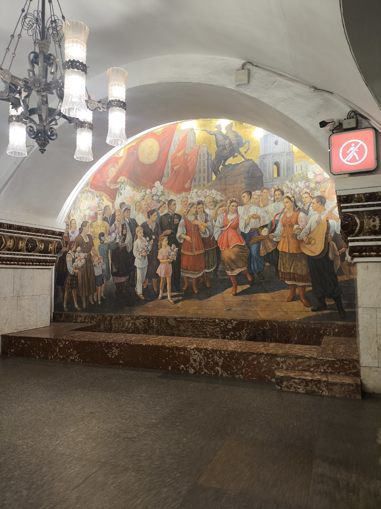 The image shows a wide mural decorating the arched wall of what appears to be a subway station. The mural depicts a large group of people, men, women, and children, in what seems to be a celebratory gathering. Many are dressed in traditional folk costumes, some holding bouquets of flowers. In the background, there are images of red banners, a cityscape with classical architecture, and a dramatic scene of a man on horseback, possibly a historical or symbolic representation. The mural is vibrant and colorful, rendered in a realistic style.
Prominently displayed on the right side of the arched ceiling is a red illuminated sign with a white symbol of a walking person crossed out, indicating "no walking" or a pedestrian prohibition. Above the mural, ornate chandeliers with frosted glass lamps hang from the ceiling, suggesting an indoor, possibly public, space. The foreground of the image shows a floor that appears to be made of dark, possibly polished stone, and a series of tiered steps made of reddish-brown marble. The overall impression is one of grandeur and cultural significance, likely a public artwork in a transportation hub. The lighting suggests it is either artificial or taken during daylight hours, as the space is well-lit. There are no visible weather cues. The emotion conveyed by the mural appears to be joyous and celebratory.
