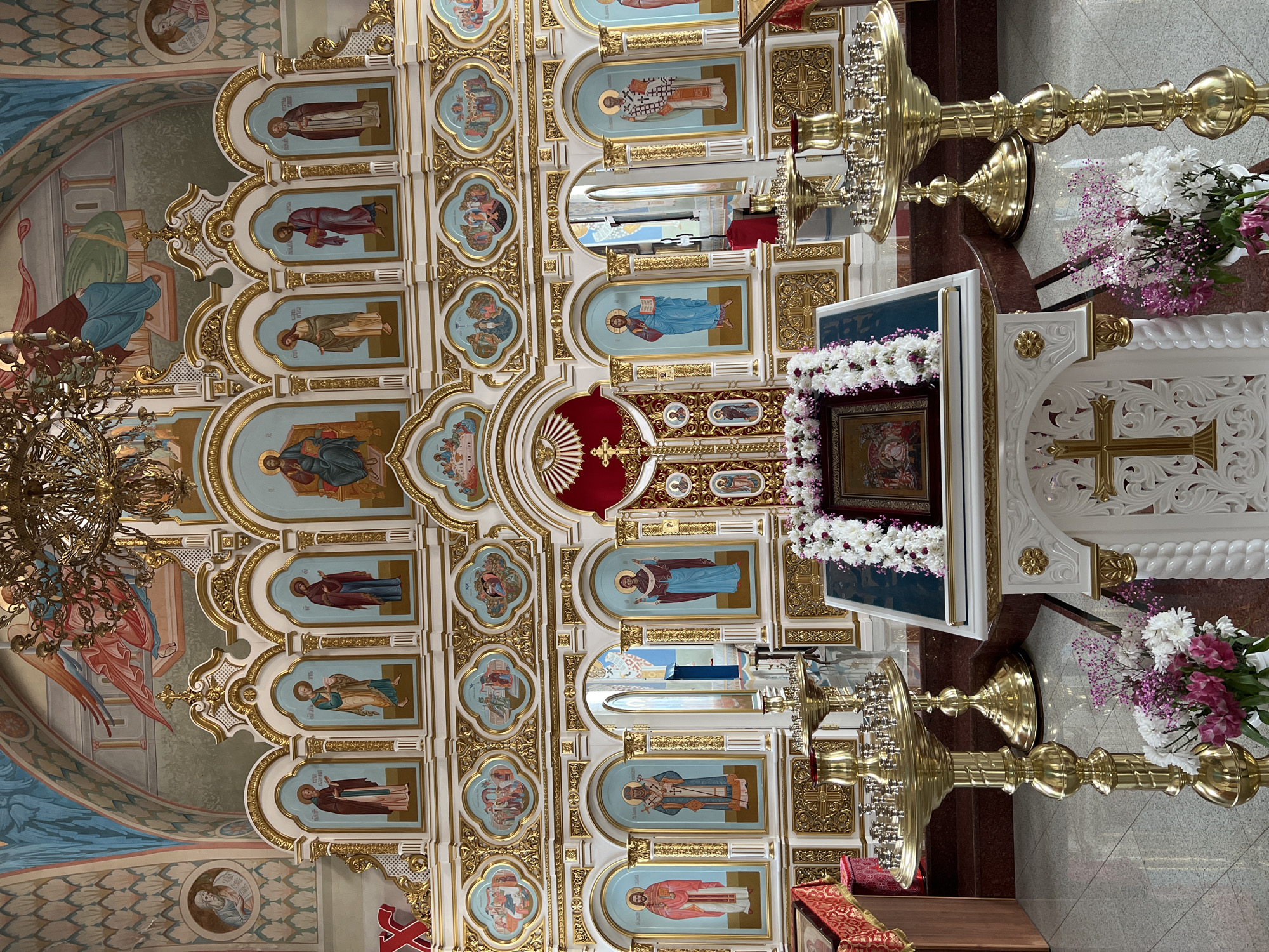The image shows the interior of a church, presumably in Kargopolye, Russia, given the prompt's context. The scene features a highly ornate iconostasis, a wall of icons and religious paintings separating the nave from the sanctuary. The iconostasis is multi-tiered with gold and white decorations. Several icons depicting religious figures are set into the wall. A lamp is suspended from the ceiling to the left. Additionally, a flower decorated stand with a religious icon is visible in front of the iconostasis. Two ornate, gold candlesticks stand on either side of the decorated stand. The floor is tiled in a light colored stone.
